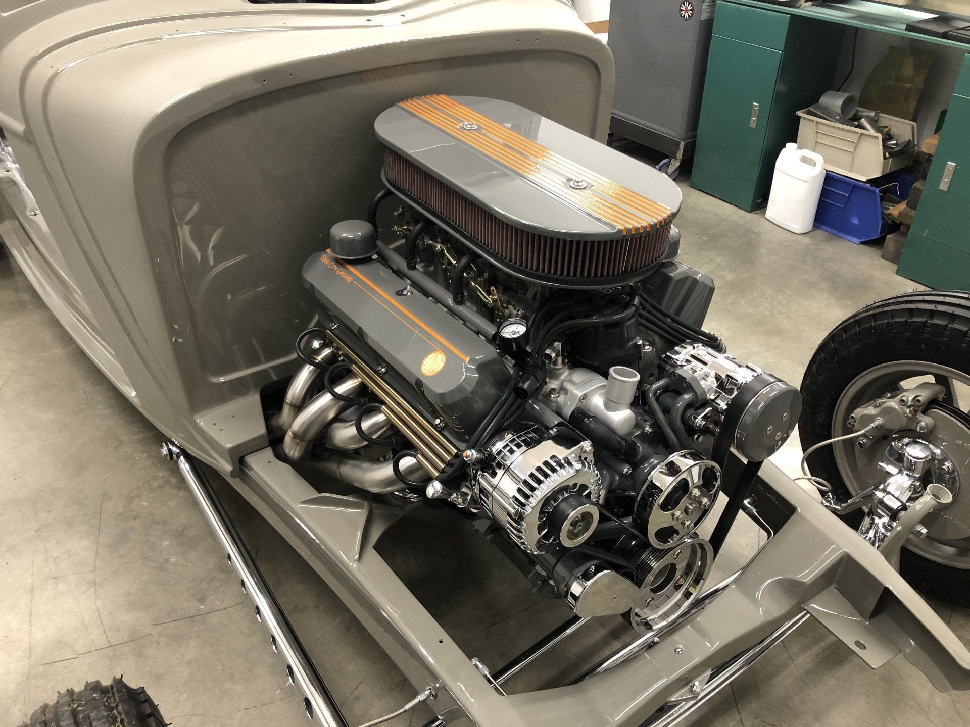 Customized car engine mounted in a gray hot rod chassis, shown in a workshop.