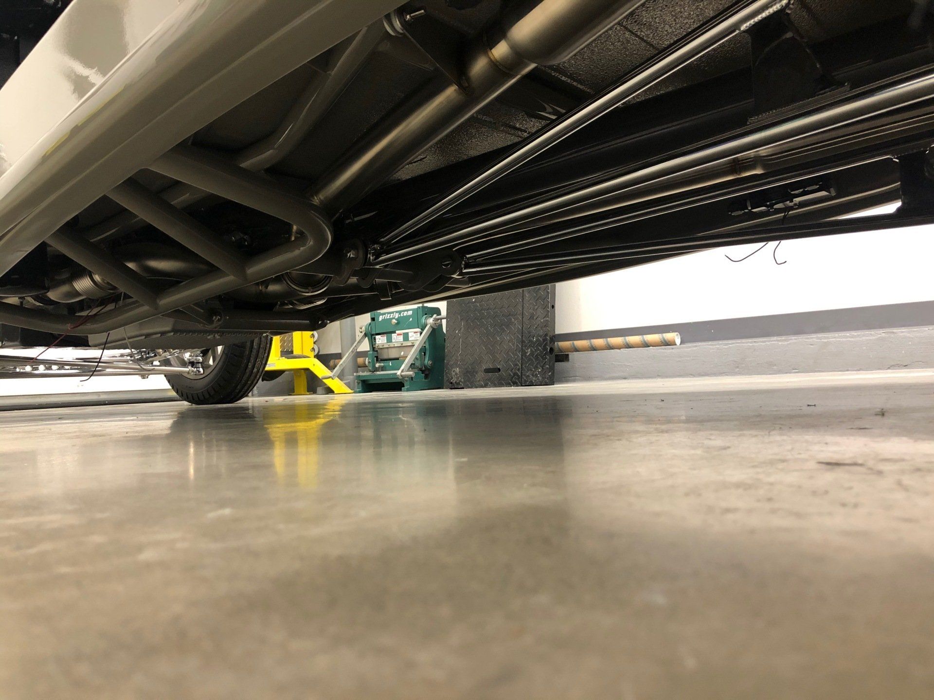 Low-angle view under a vehicle on a garage floor, showing metal pipes, a yellow jack stand, and a green tool cart.