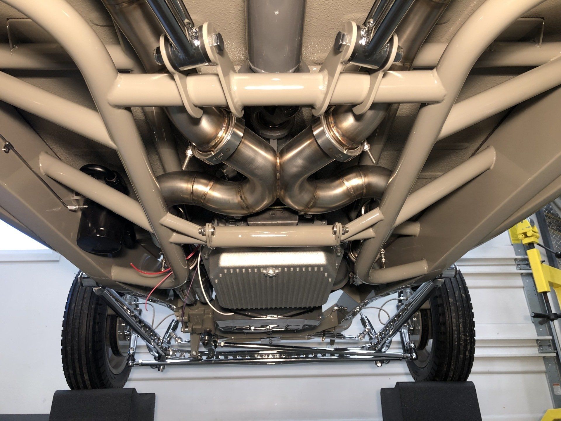 Underside of a vehicle showing exposed chassis, exhaust piping, and suspension components in a garage.