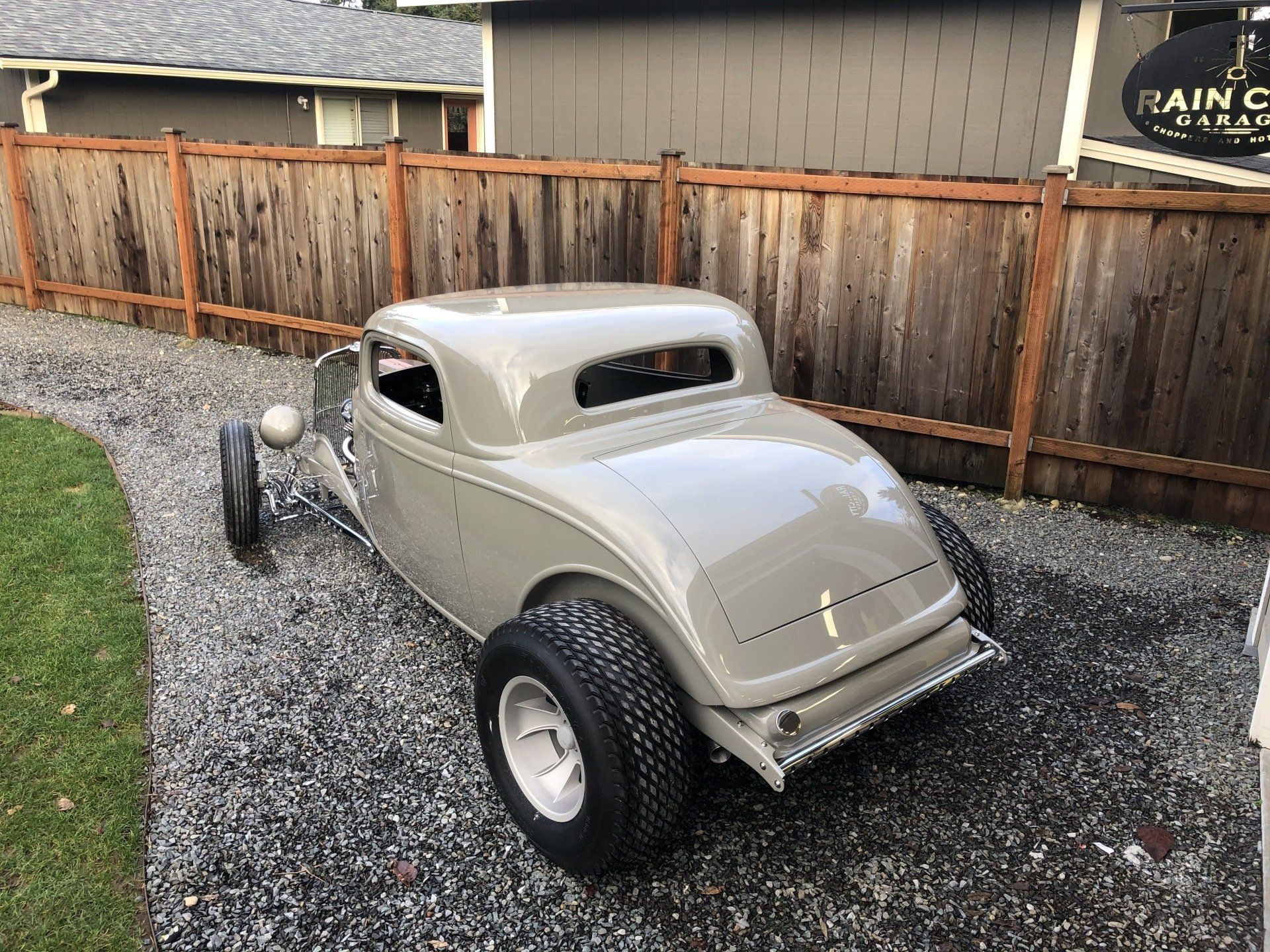 Silver vintage hot rod parked on gravel beside a wooden fence in a backyard.