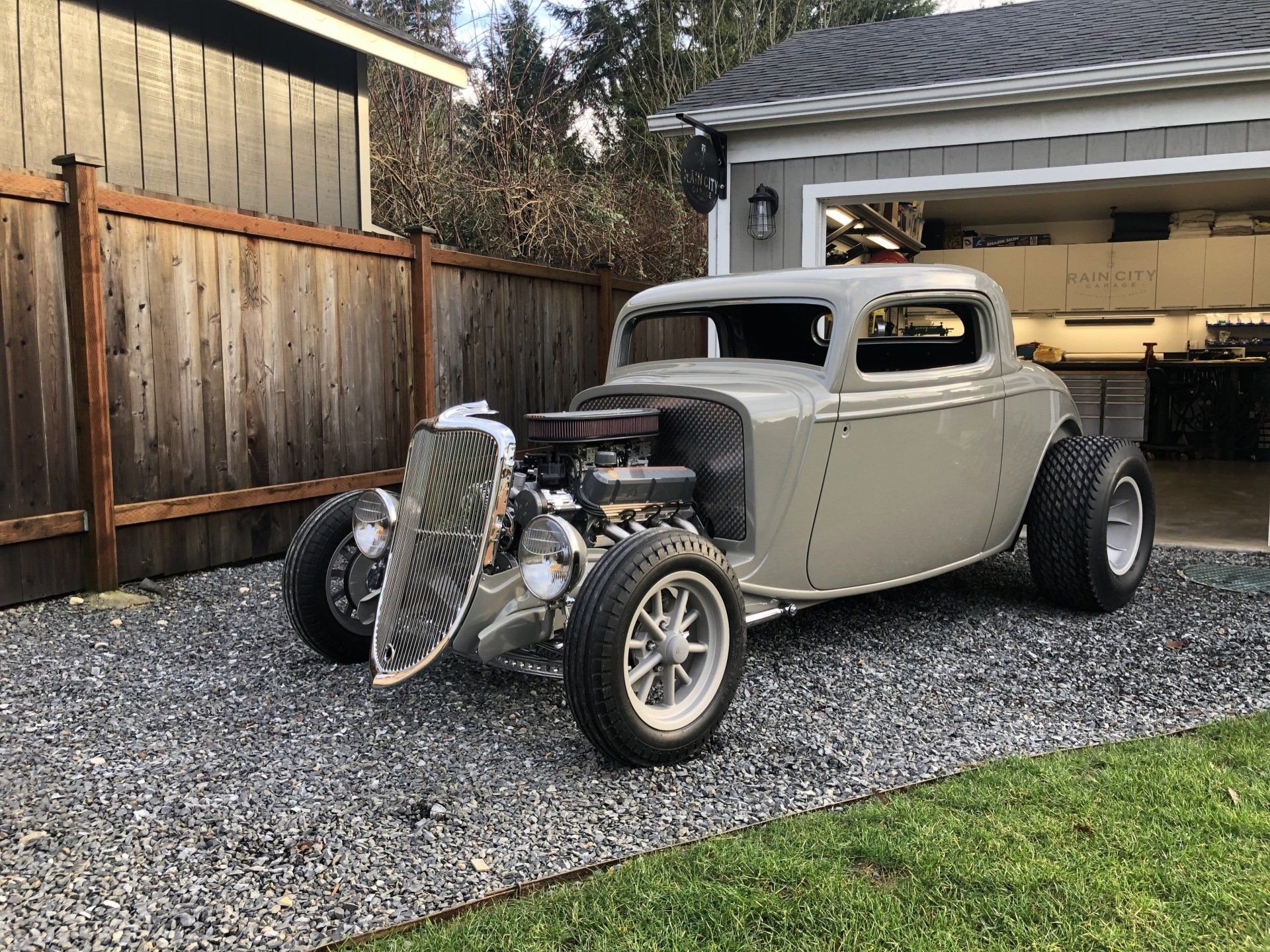 Vintage gray hot rod parked on gravel driveway beside a wooden fence and open garage
