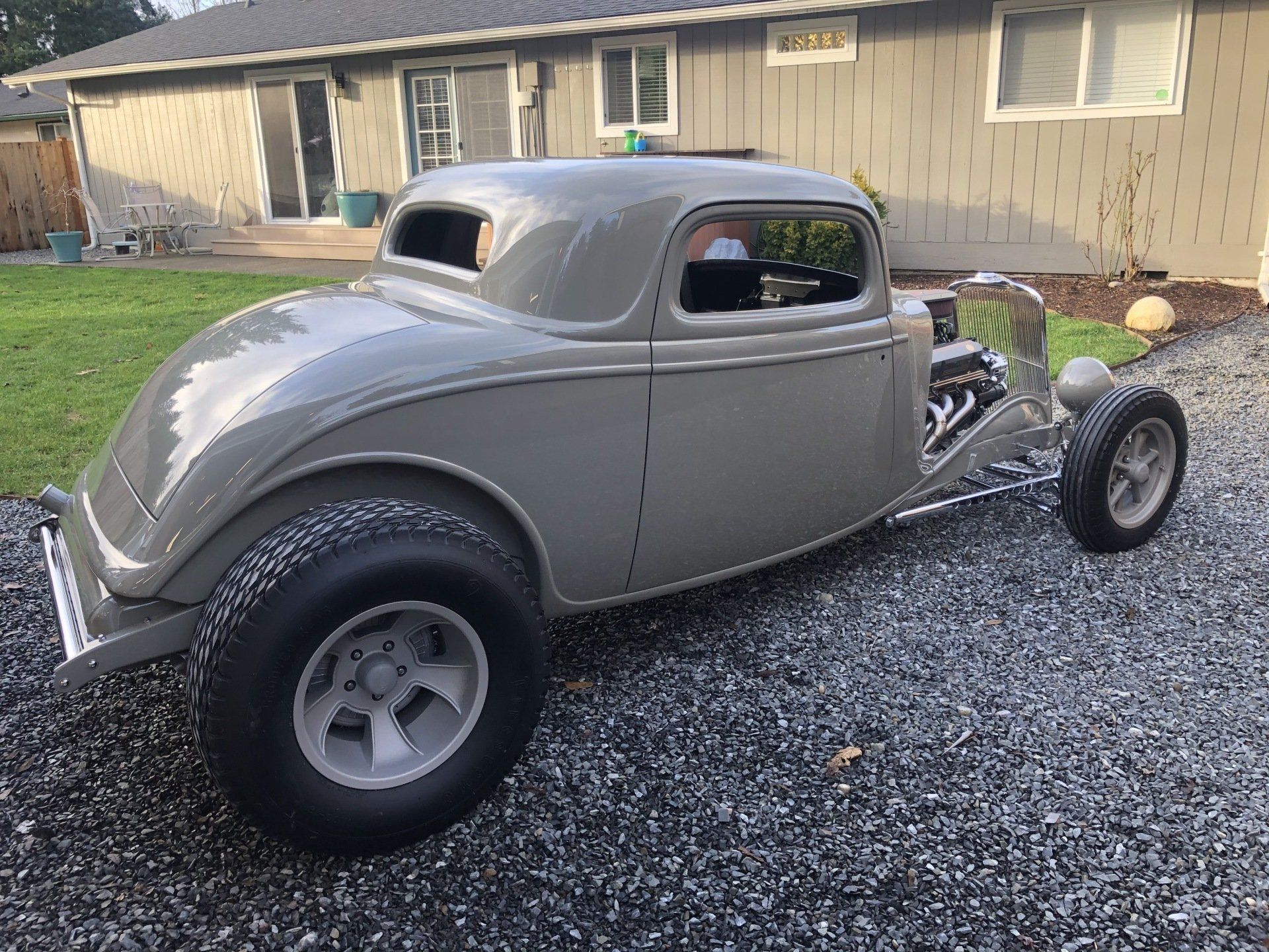 Gray hot rod coupe parked on gravel in front of a beige house, shown in side view.
