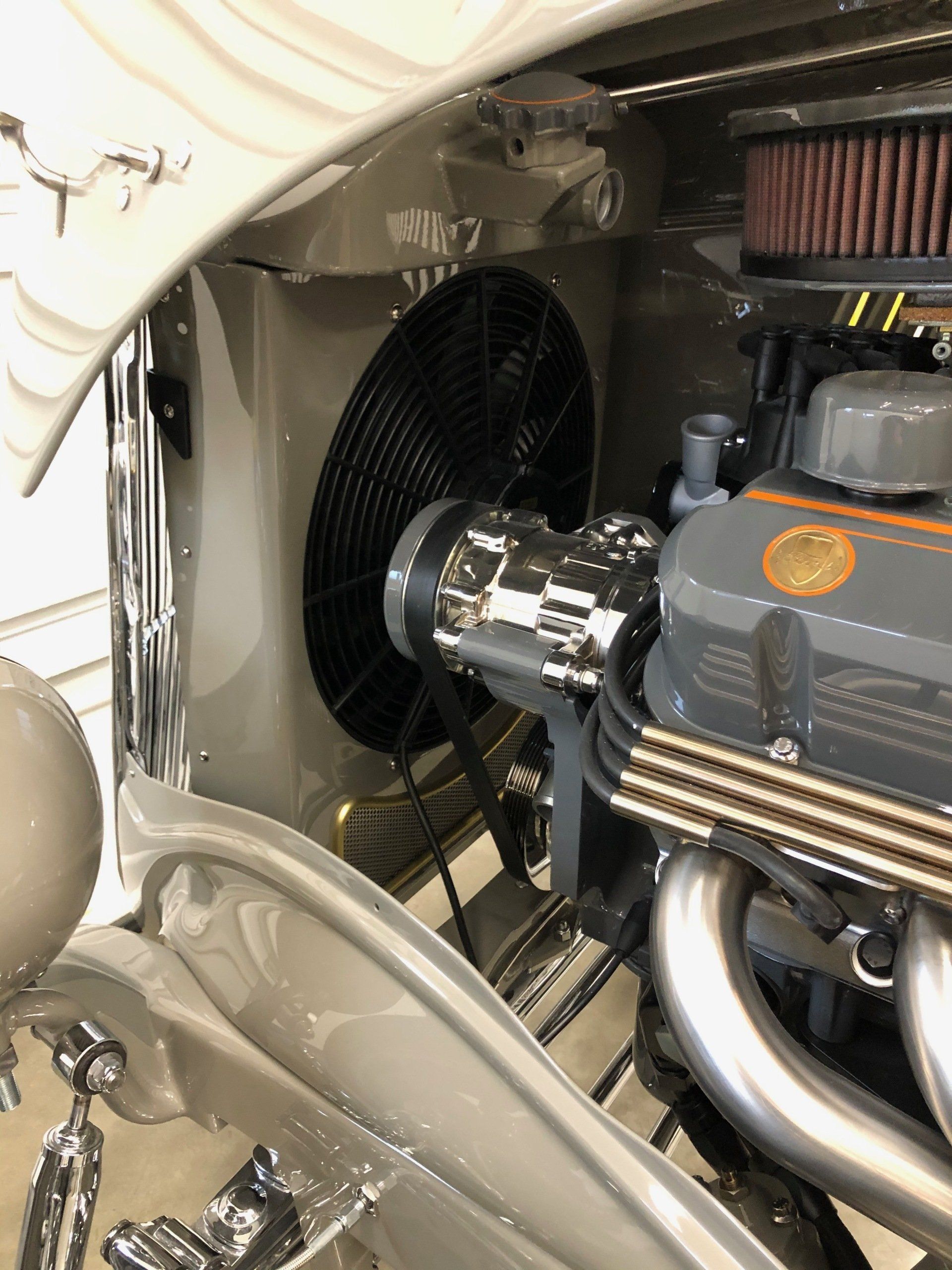 Classic car engine bay with chrome intake, polished headers, and a black cooling fan.