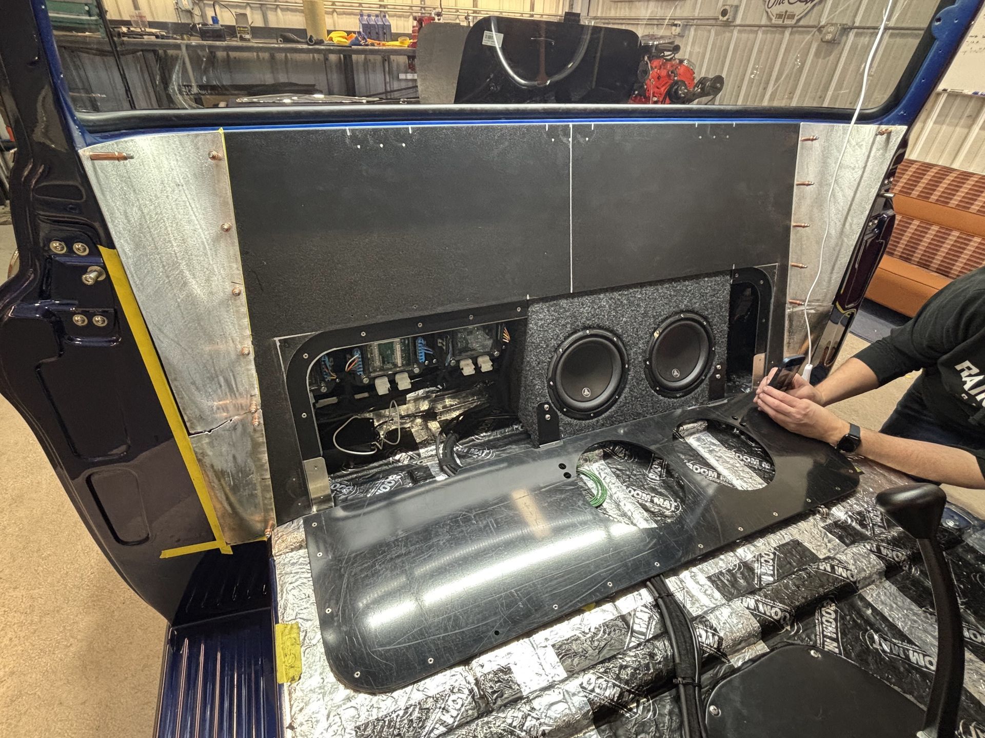 Car audio display with exposed subwoofer and wiring, black panels, and a person crouching beside it.