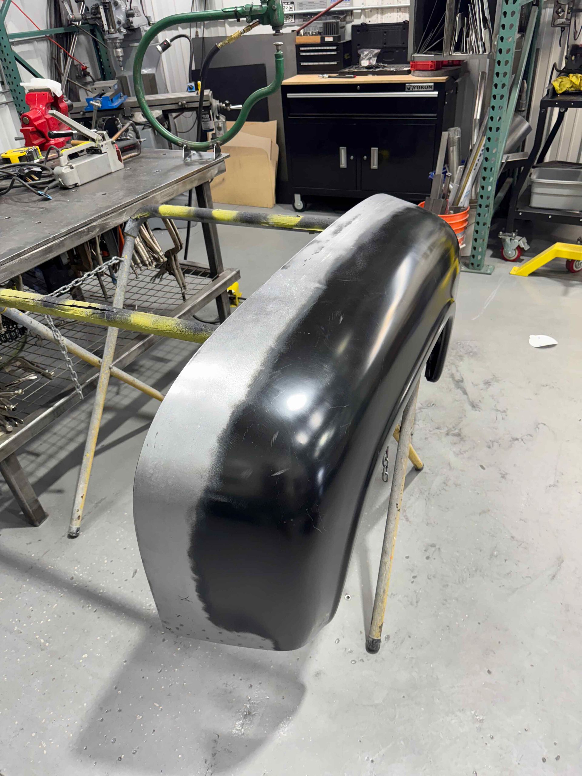 Glossy black car bumper or body panel on stands in a workshop, with tools and equipment around it