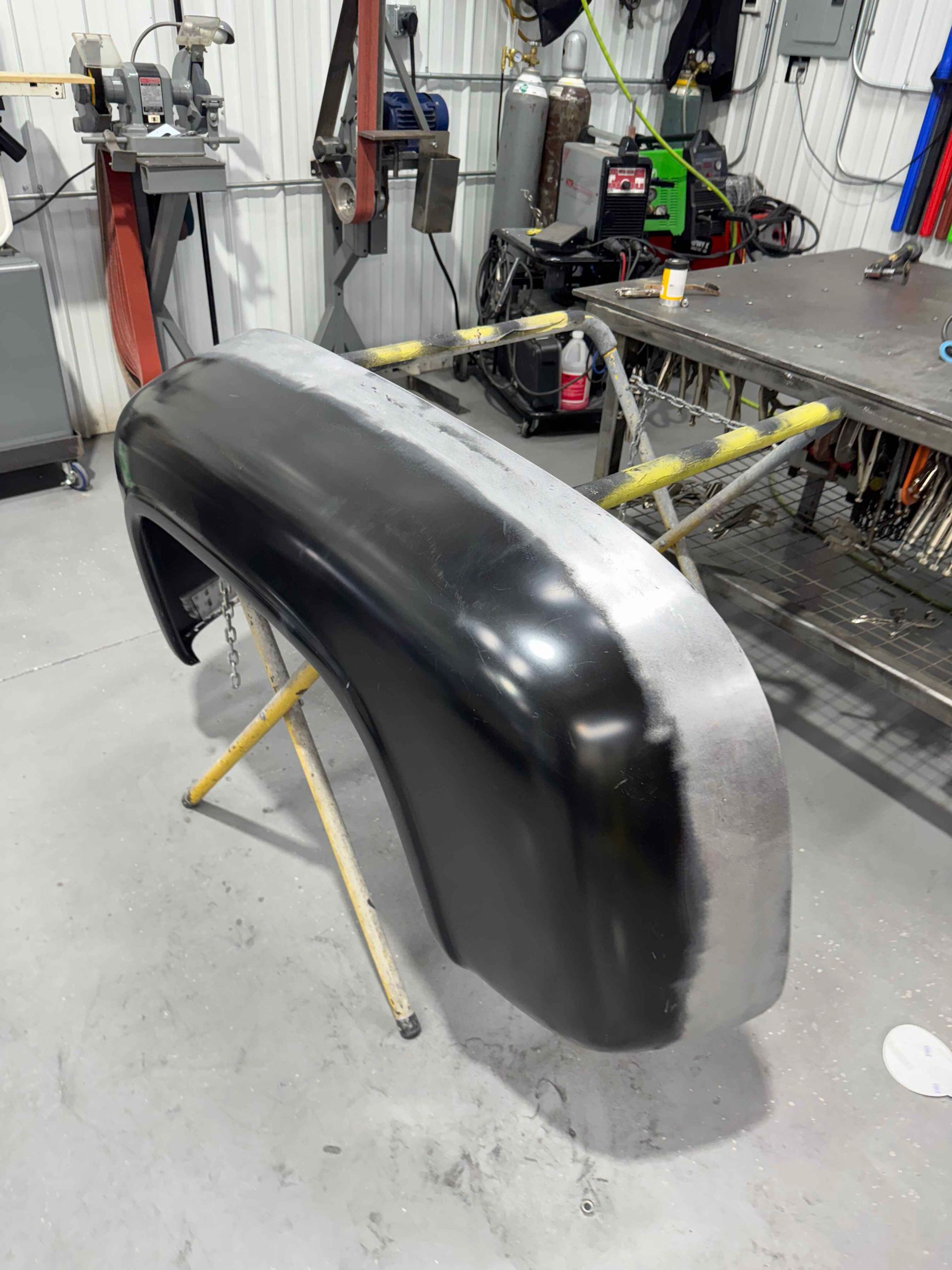 Black car body panel on a stand in an auto workshop, with tools and equipment in the background