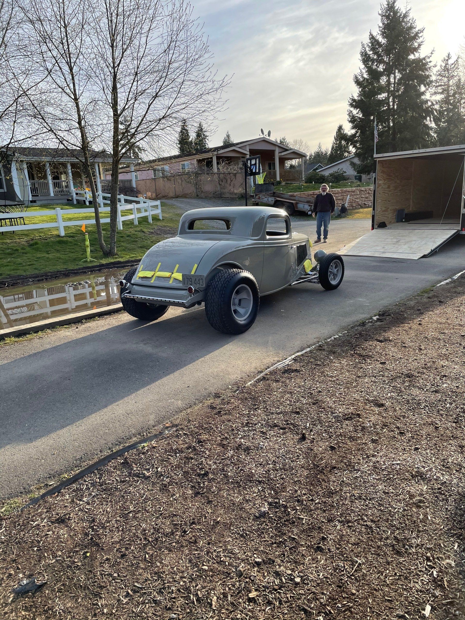 Vintage gray hot rod with neon yellow accents parked on a driveway beside a gravel path, two people nearby.