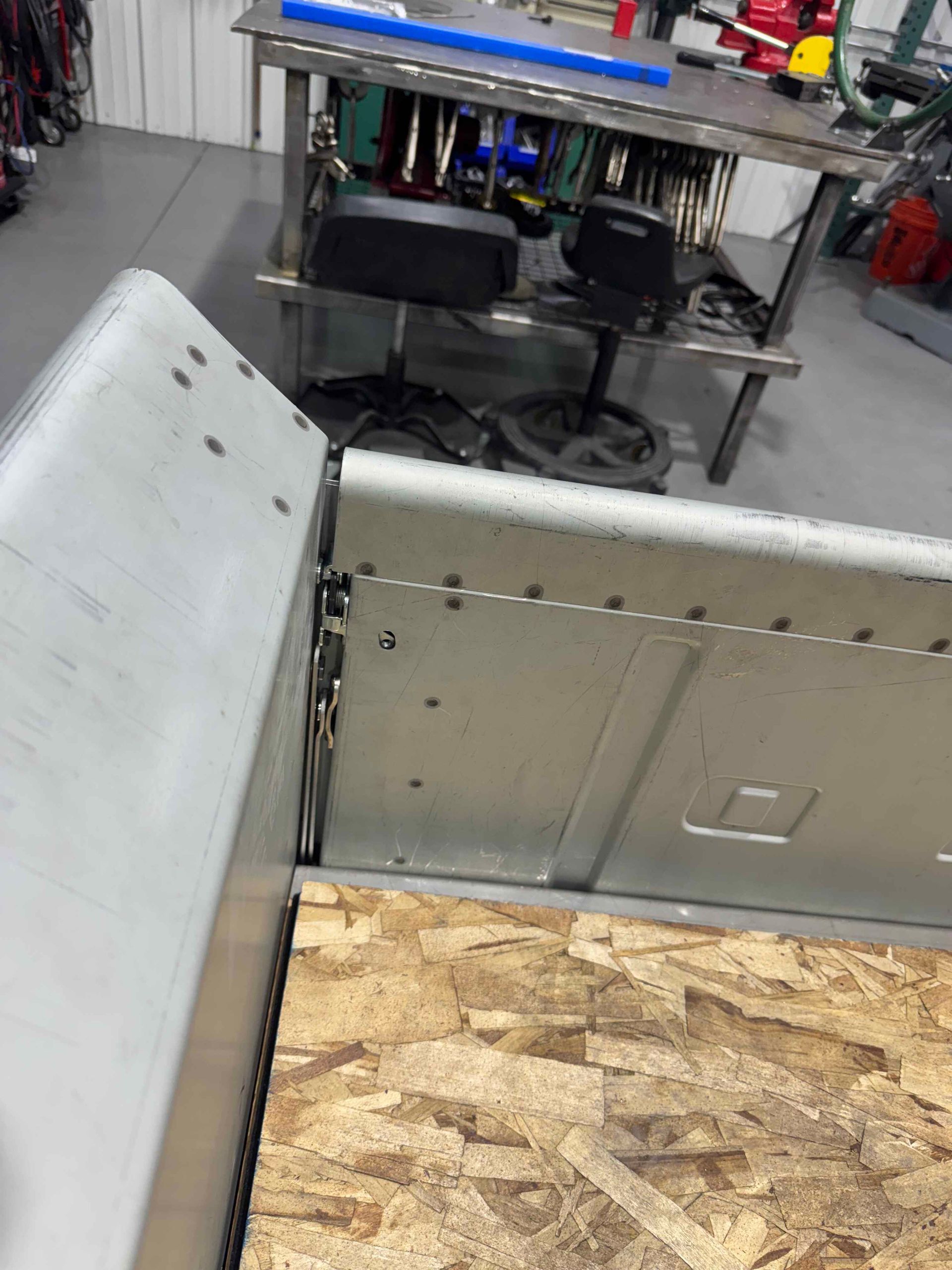 Open storage lid in a workshop, showing plywood interior and a cluttered workbench behind it
