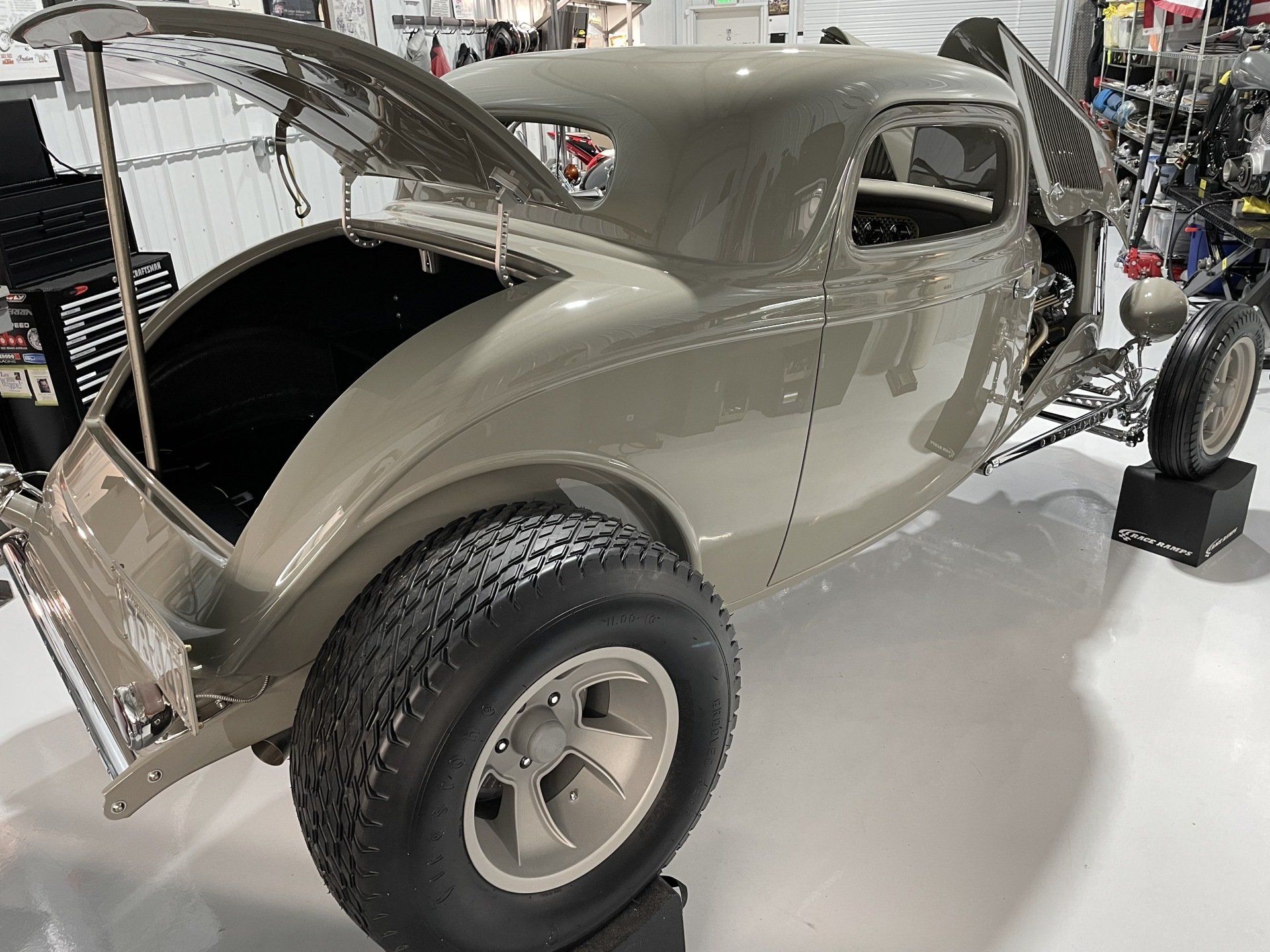 Vintage beige hot rod coupe with rear hatch open, displayed indoors on a showroom floor.