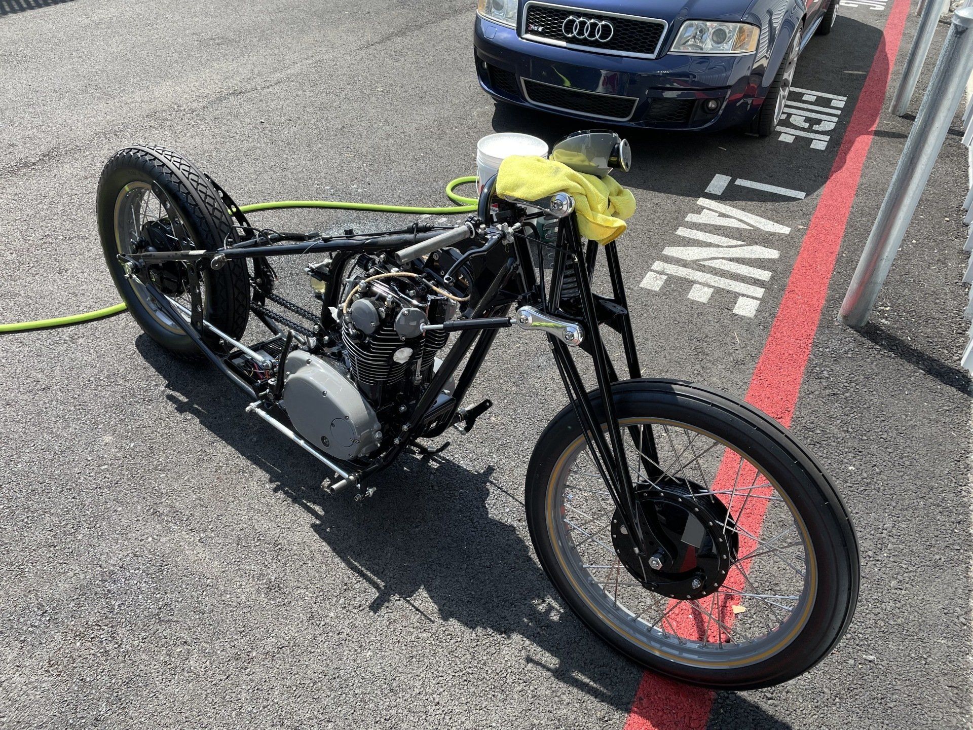 Custom black motorcycle parked on pavement with a yellow bag on the seat, near a curb and parked car