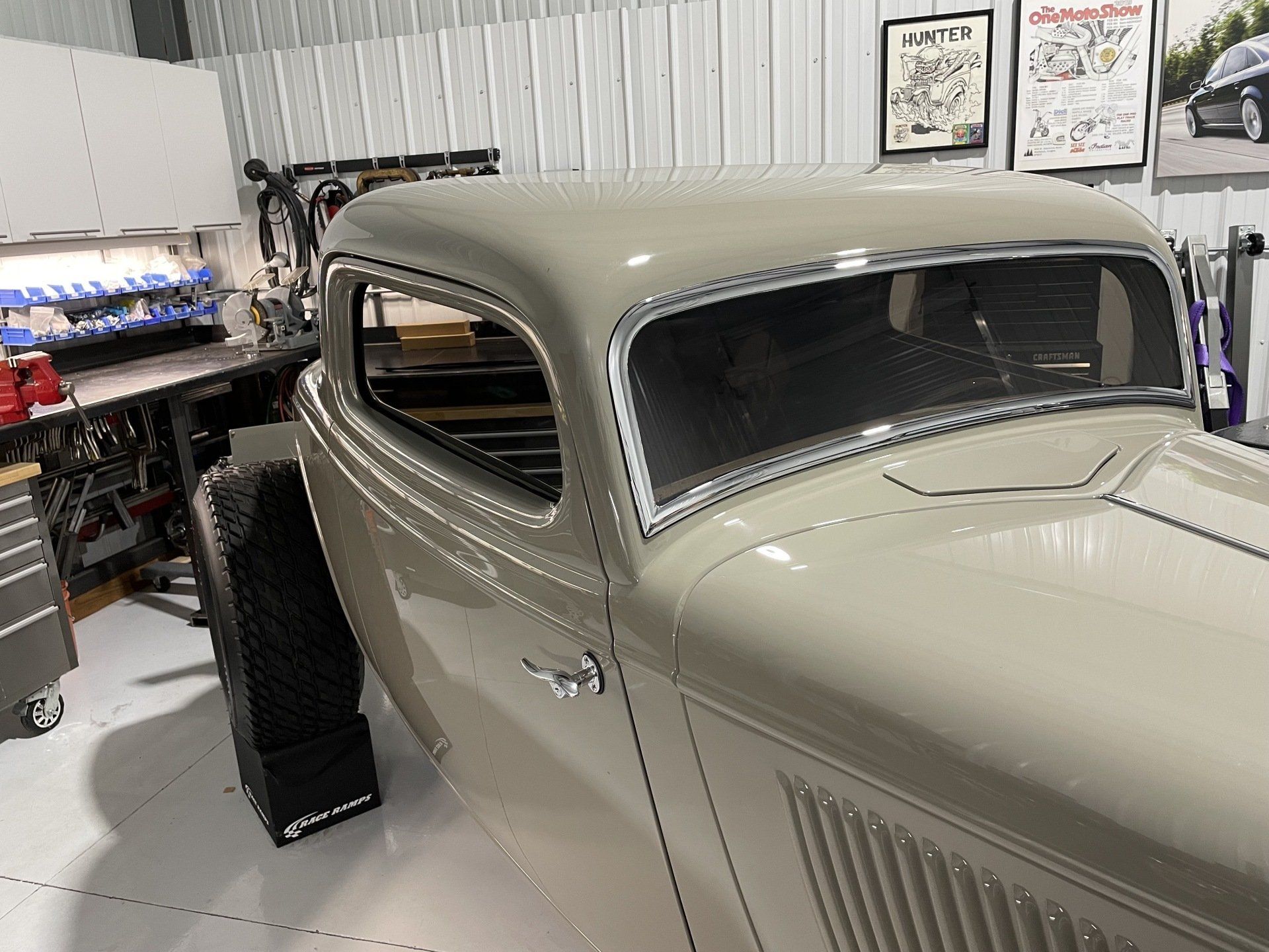 Vintage gray car in a workshop, shown from the front side beside tools and wall art.