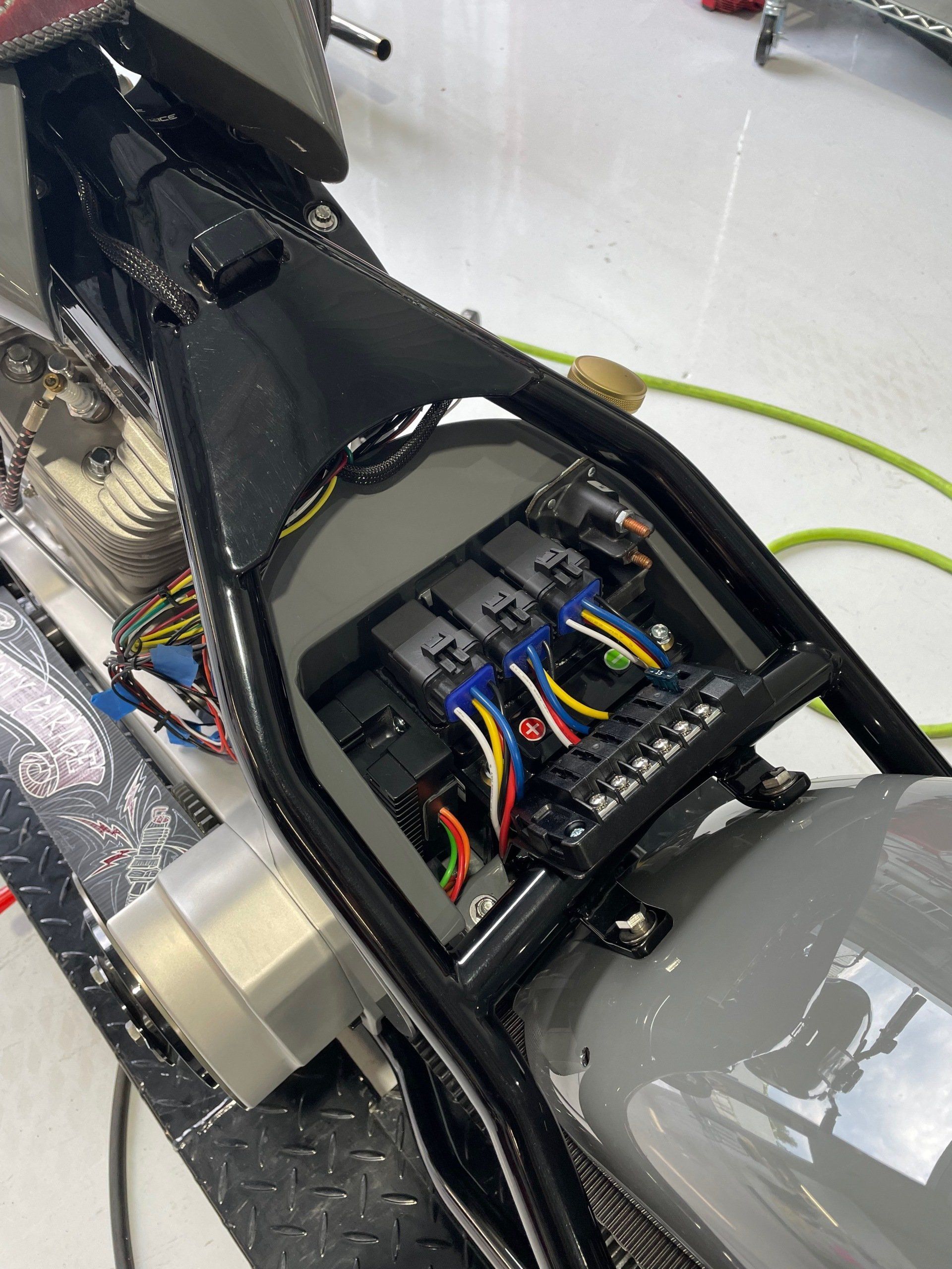 Open motorcycle seat with exposed wiring and electronics, showing a black frame and gray bodywork.