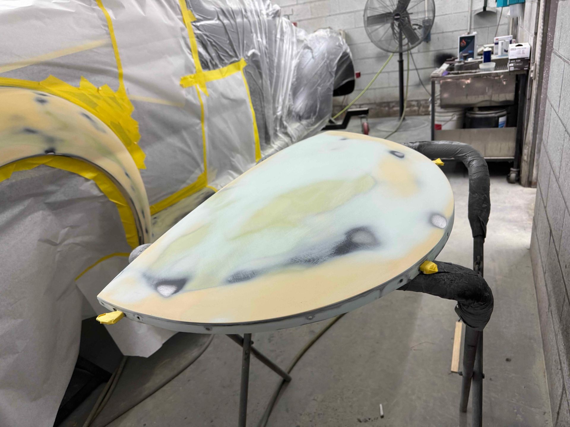 Surfboard with primer and yellow masking in a workshop, set on a stand beside covered equipment.