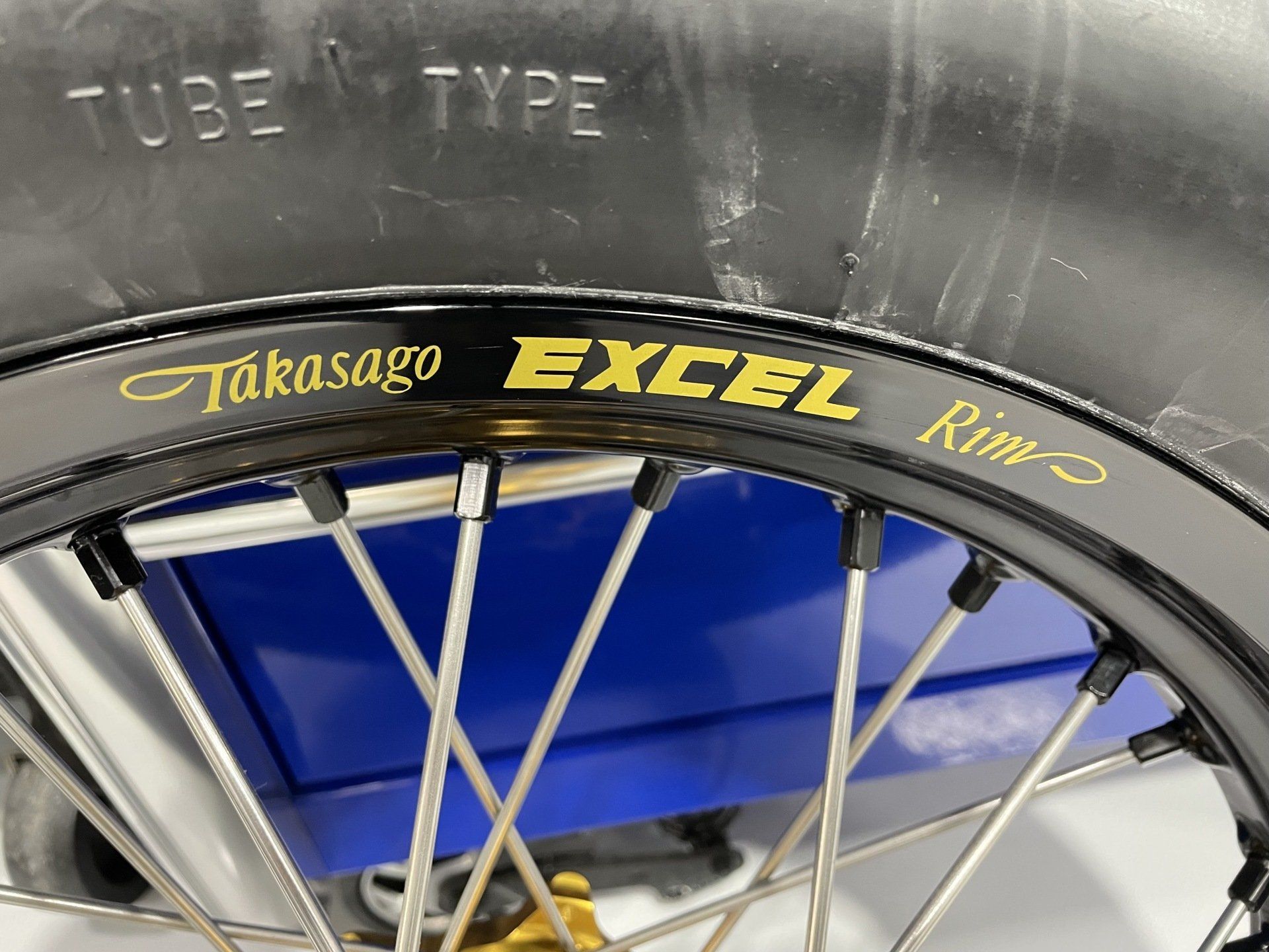 Close-up of a bicycle wheel with a black tire labeled Takasago EXCEL on a blue background