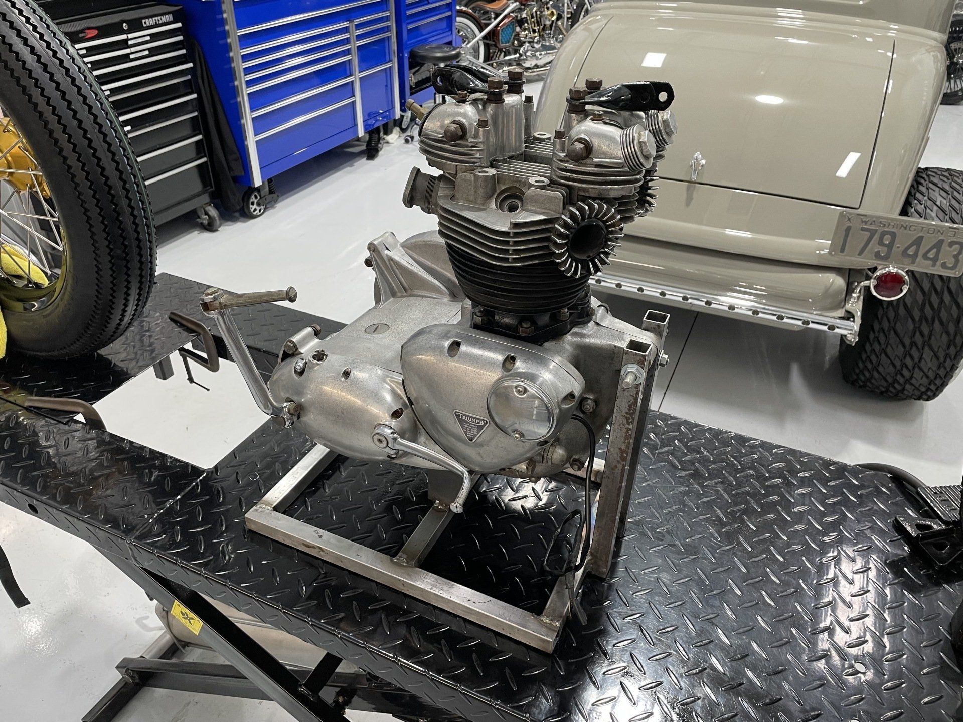 Small motorcycle engine mounted on a metal frame in a workshop, with a tire and tool carts nearby