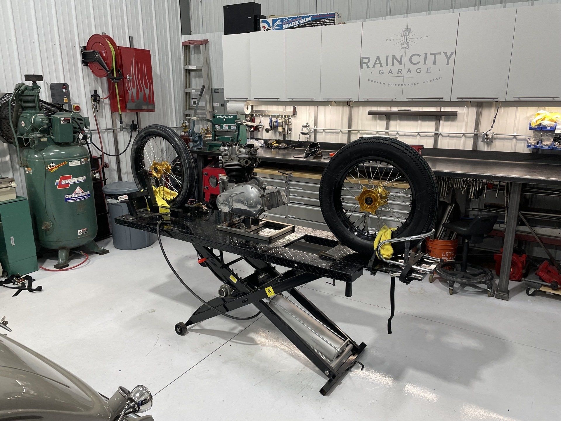 Motorcycle frame on a lift in a workshop, with tools, tires, and green gas cylinders nearby.