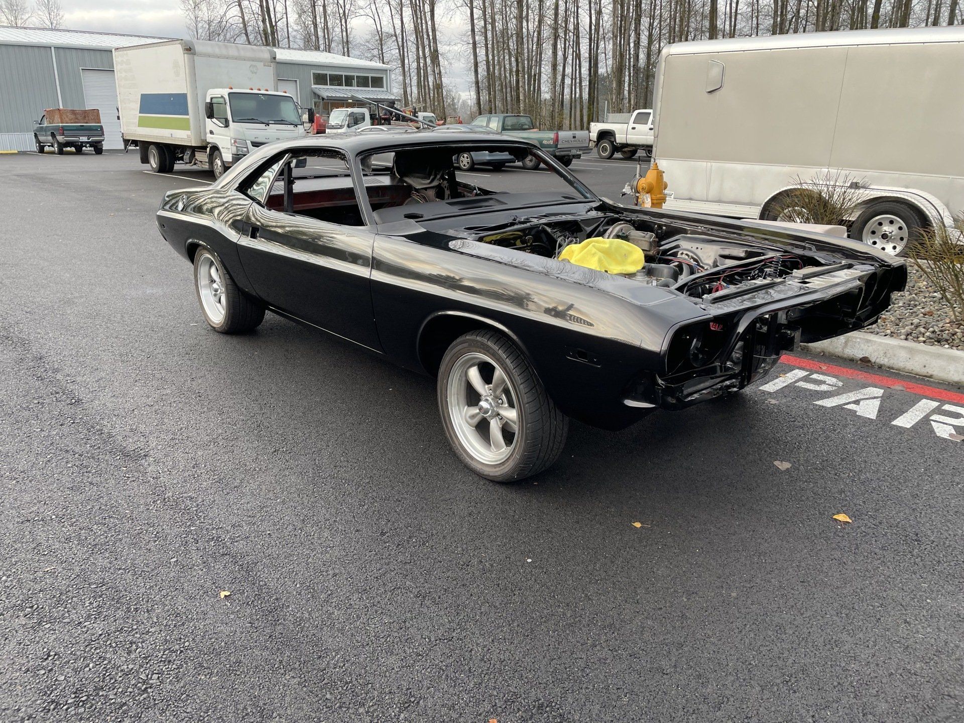 Black classic car with exposed engine parked on wet lot, with trucks and trailer in background