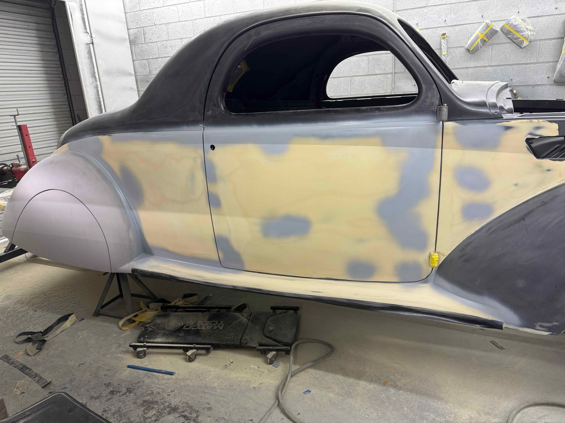 Classic car body in a garage, partially sanded and primed with gray and yellow body filler.