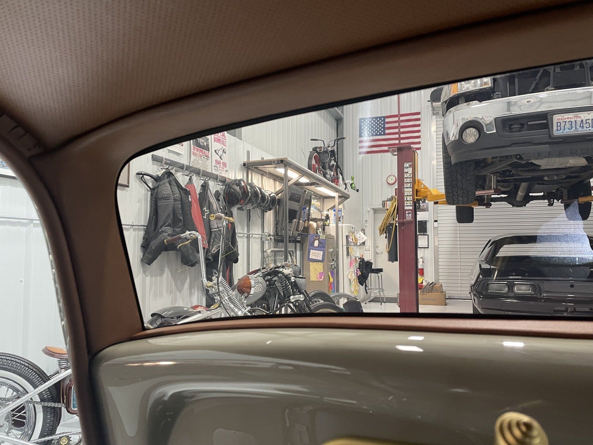 Inside a garage, a vintage car interior looks out toward a workshop with hanging tools and a lifted truck.