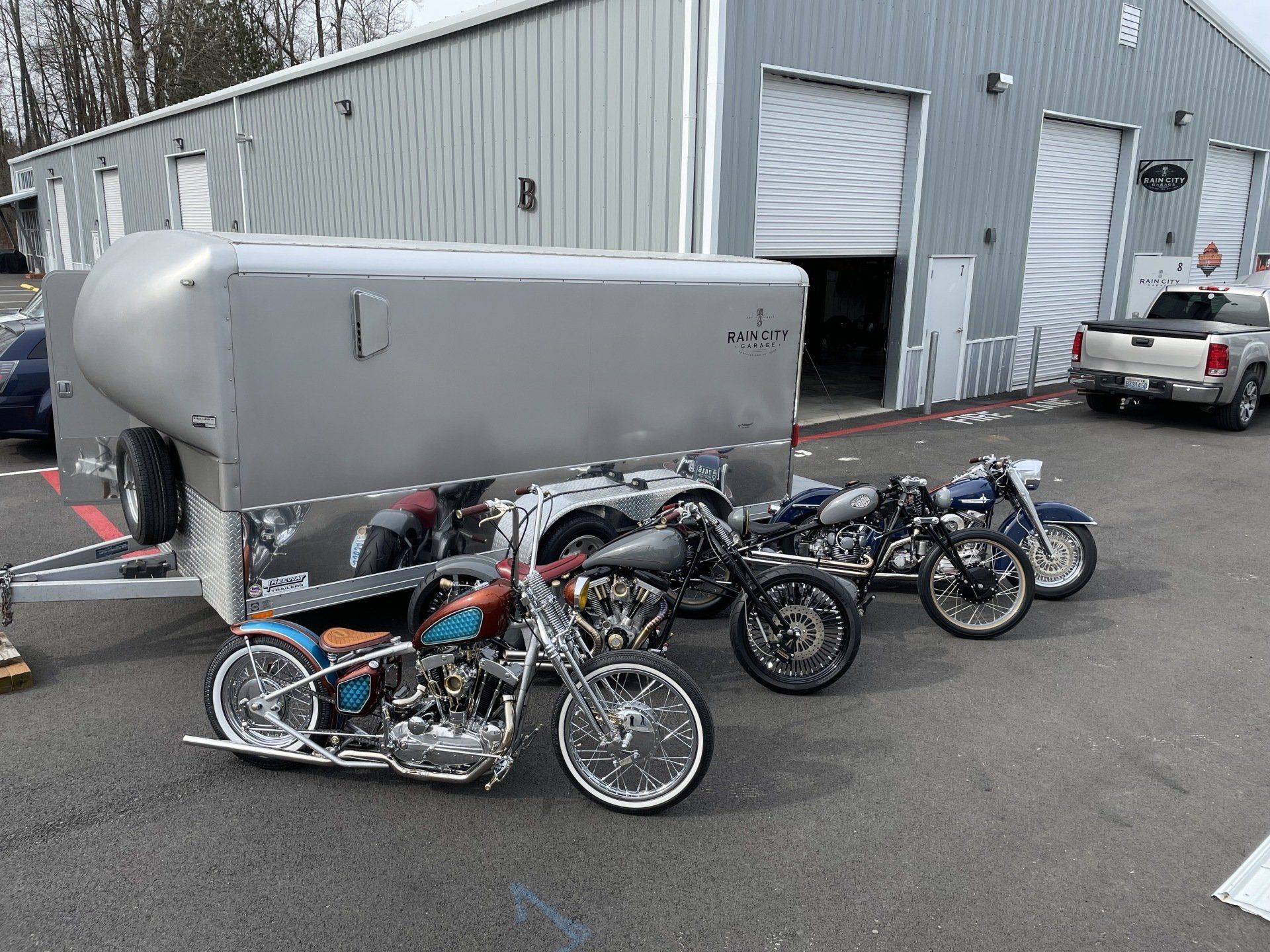 Four custom motorcycles parked outside a gray industrial building with white garage doors