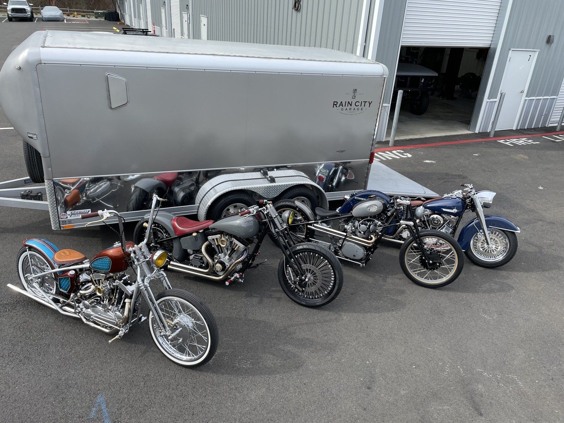 Four custom motorcycles parked beside a gray enclosed trailer in an industrial lot.