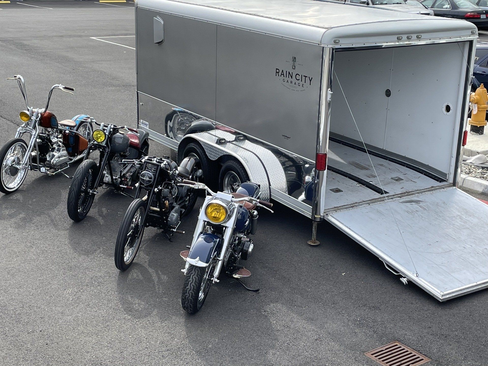 Three motorcycles parked beside an open gray trailer on a paved lot