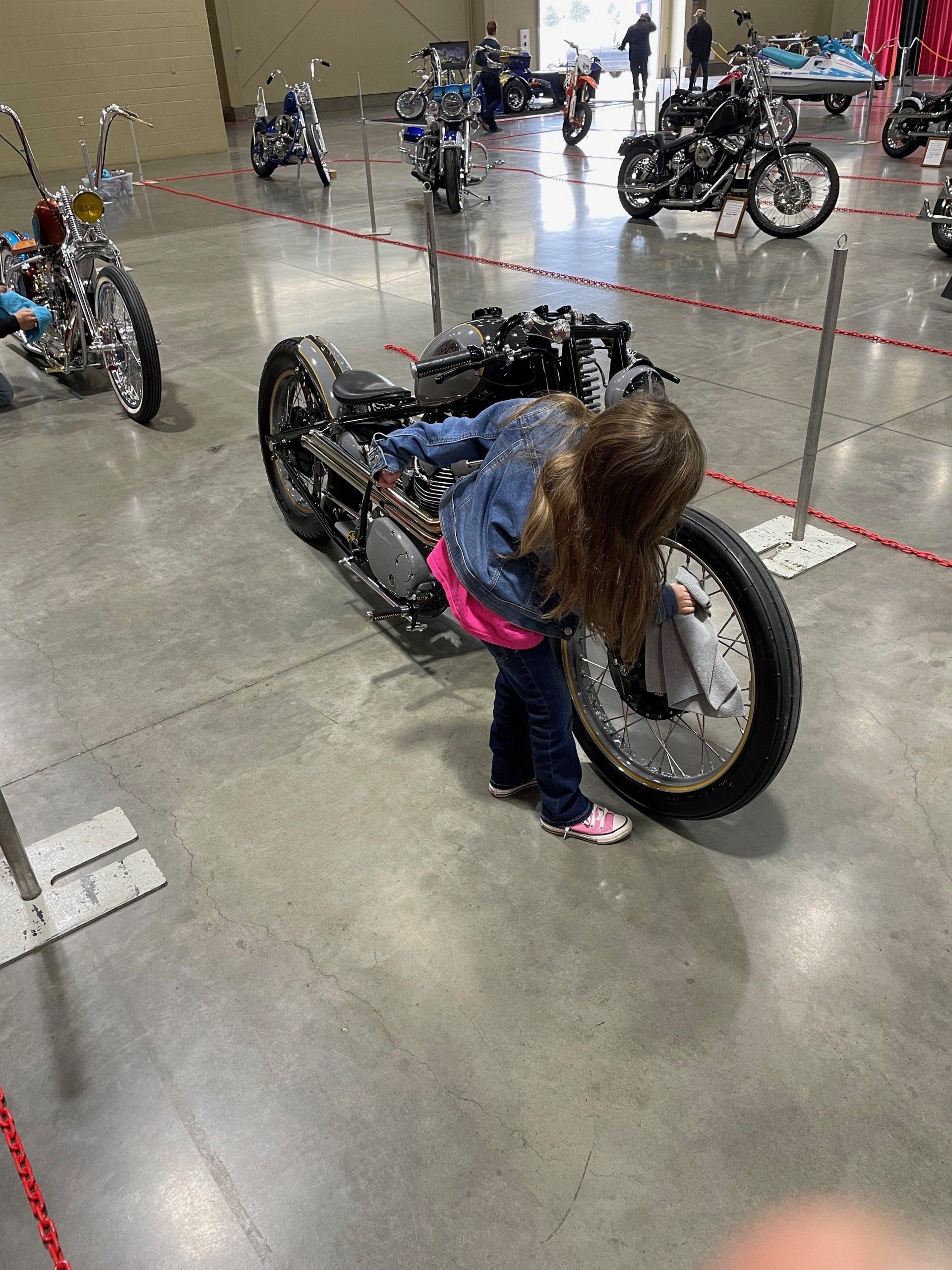 Child crouching beside a black motorcycle in a large indoor showroom or event space.