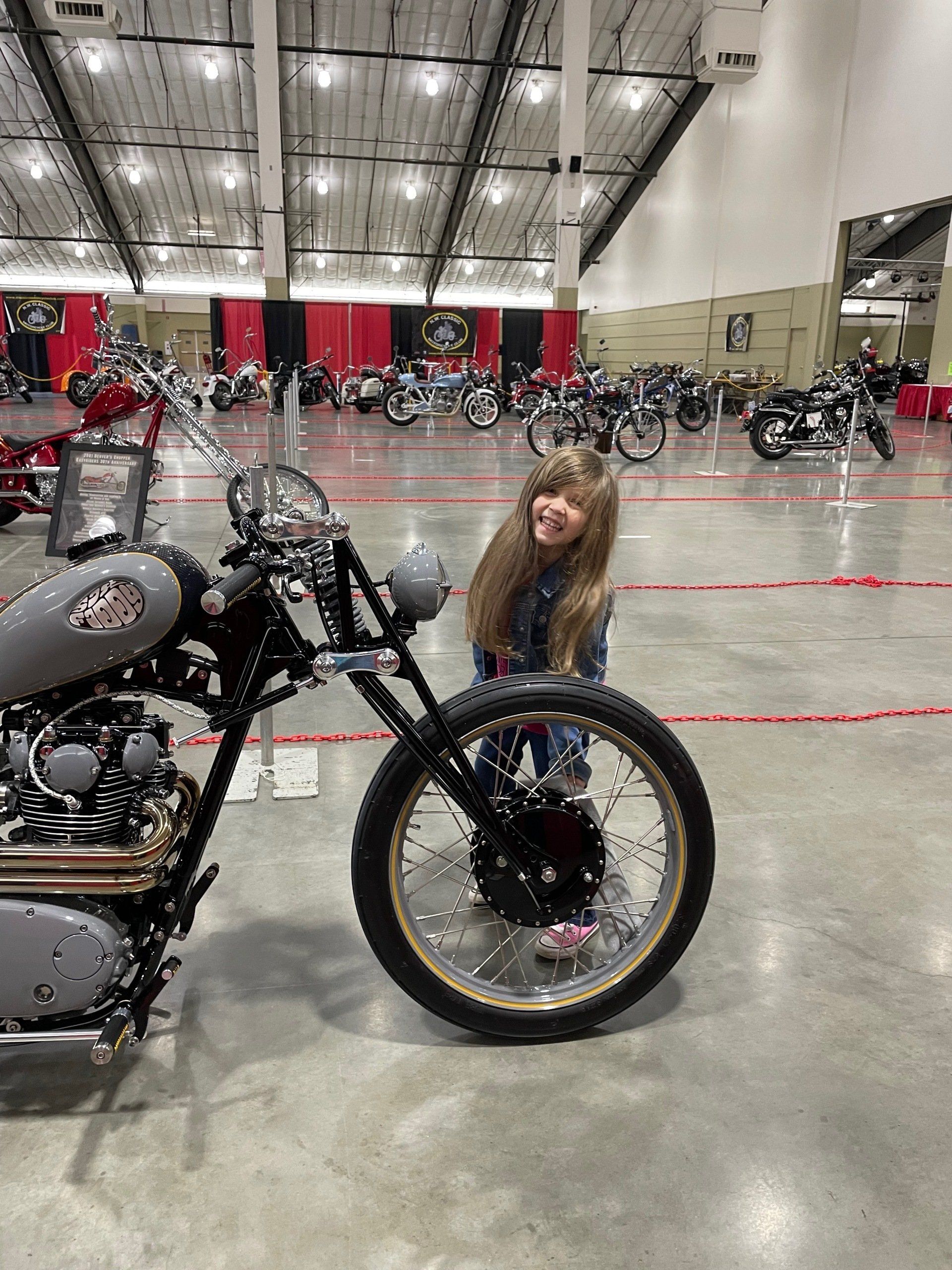 Woman standing beside a black motorcycle in a large indoor convention hall with rows of bikes in the background