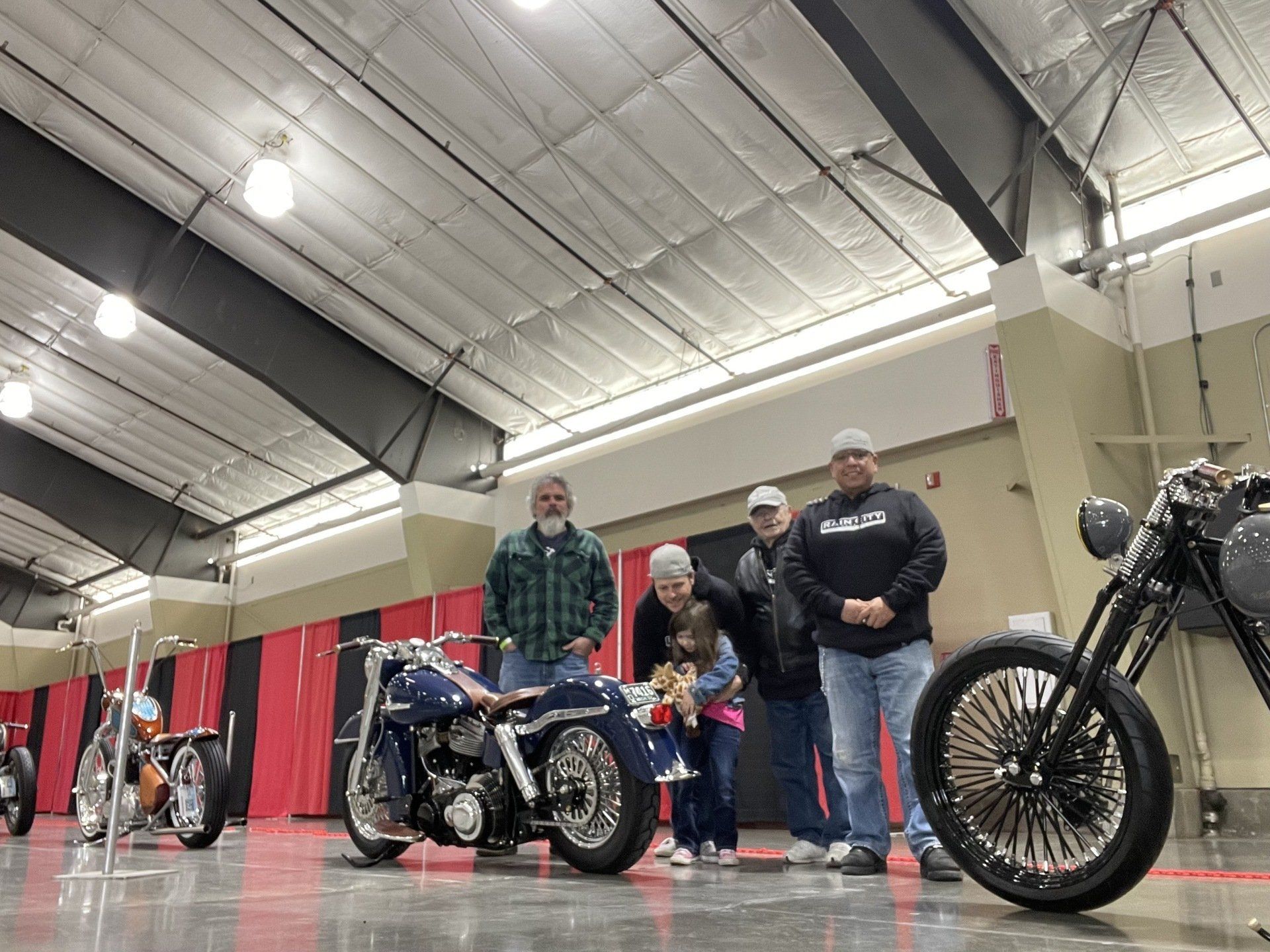 Three people stand beside custom motorcycles at an indoor bike show.