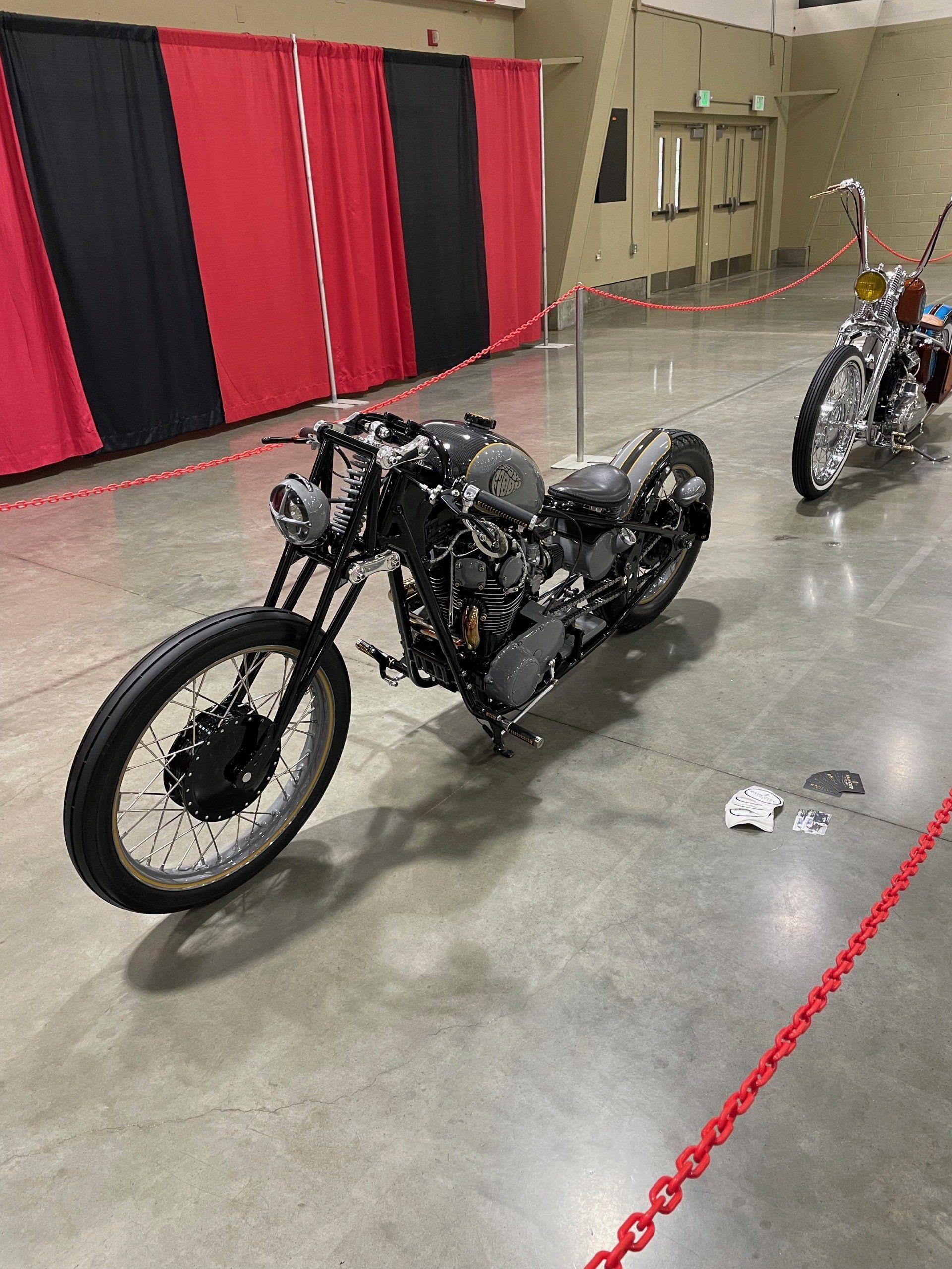 Black motorcycle on display indoors at an exhibition, with a second bike in the background.