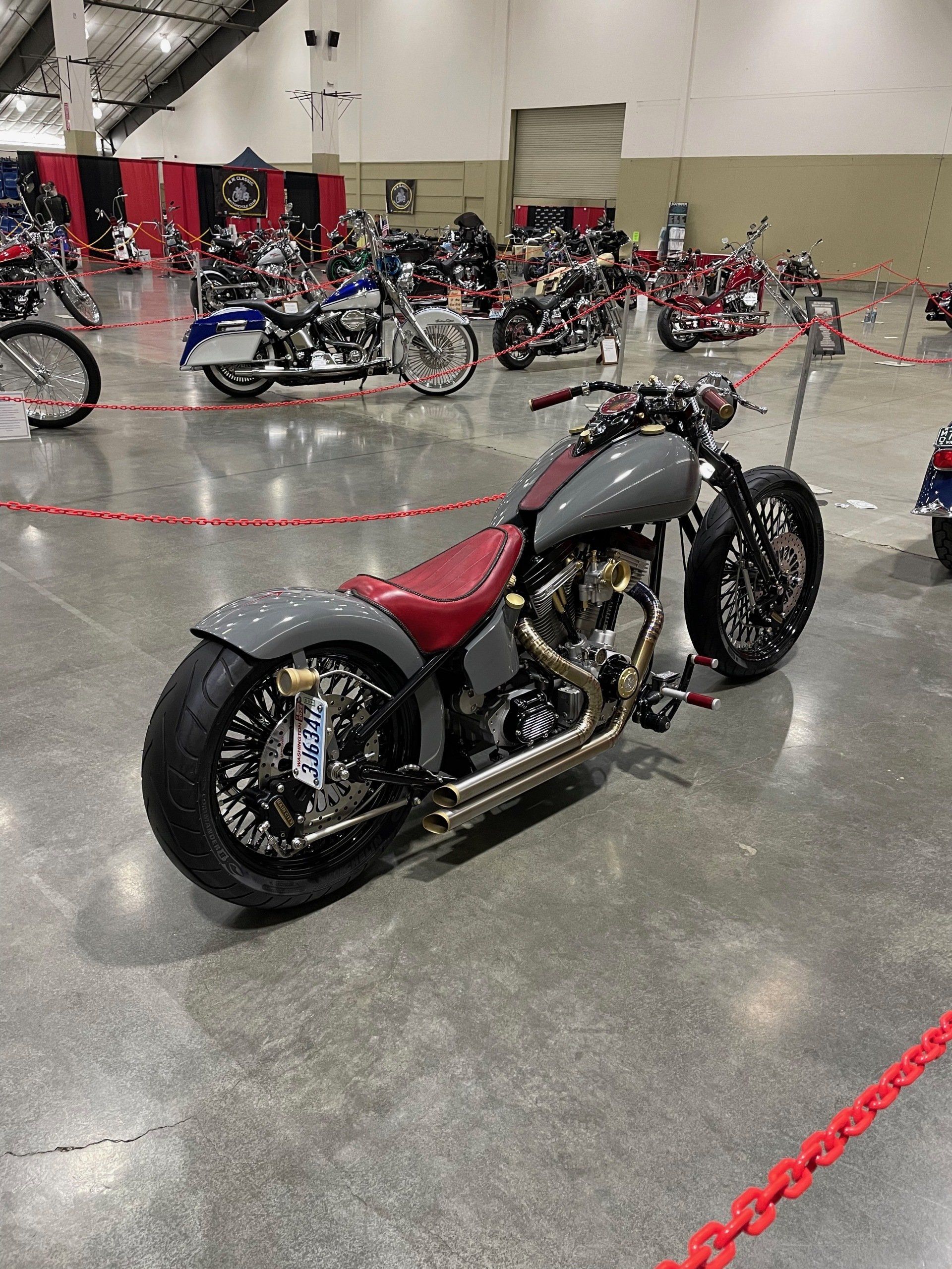 Custom motorcycle with red seat on display in an indoor showroom, surrounded by other bikes and red barrier ropes