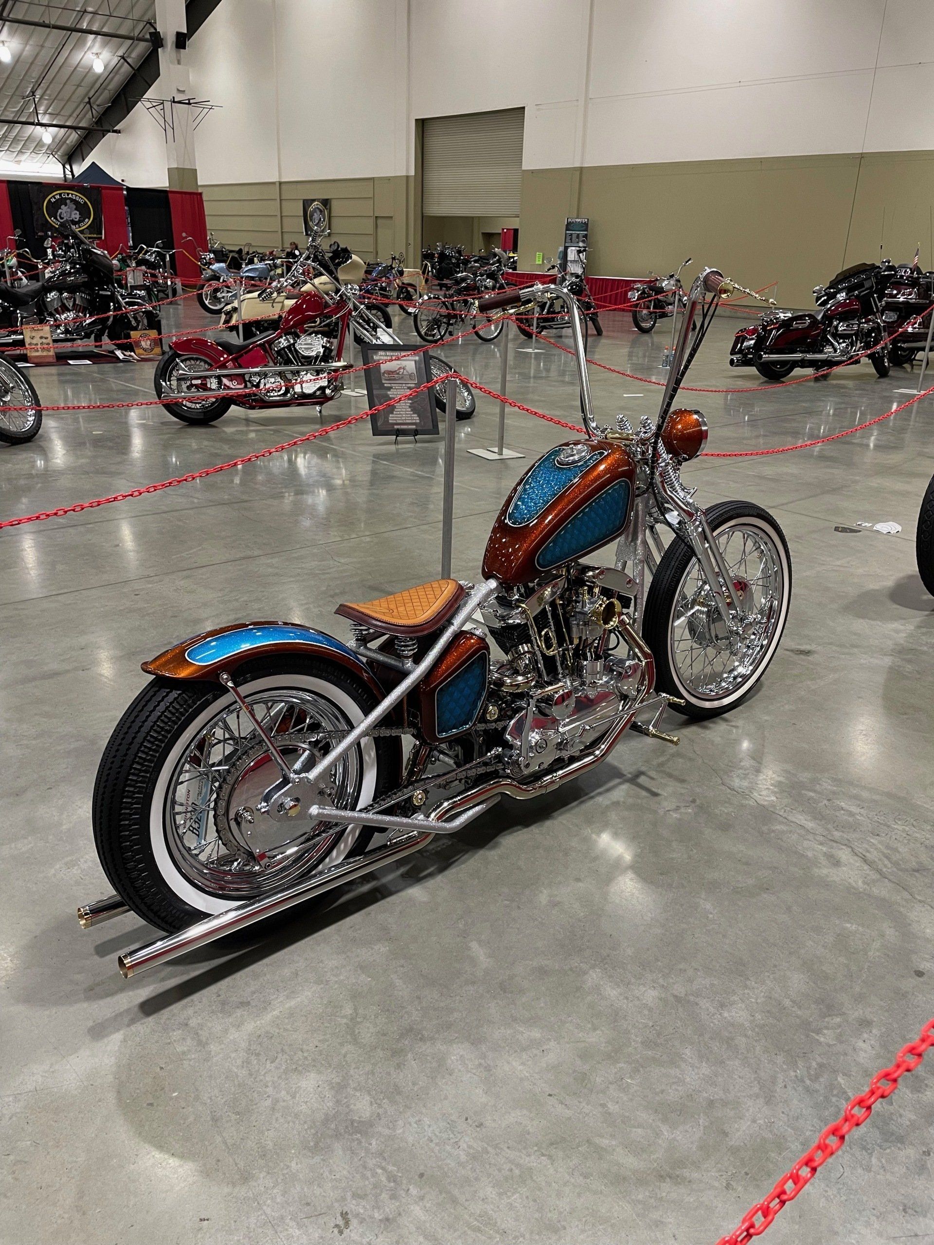 Custom orange-and-blue motorcycle displayed indoors on a polished floor, with other bikes in the background.