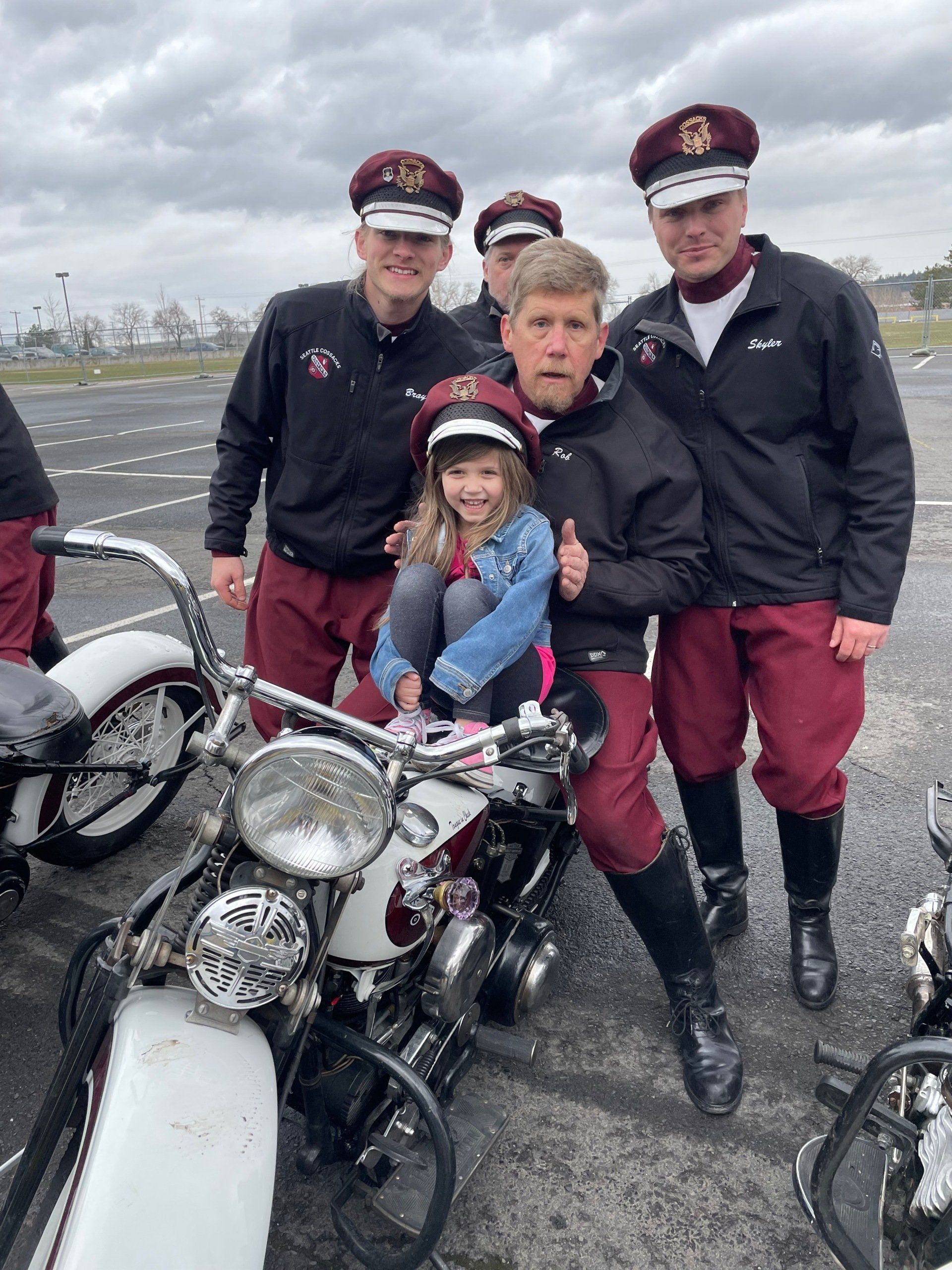 Person and child on a motorcycle with uniformed riders in the background on a runway