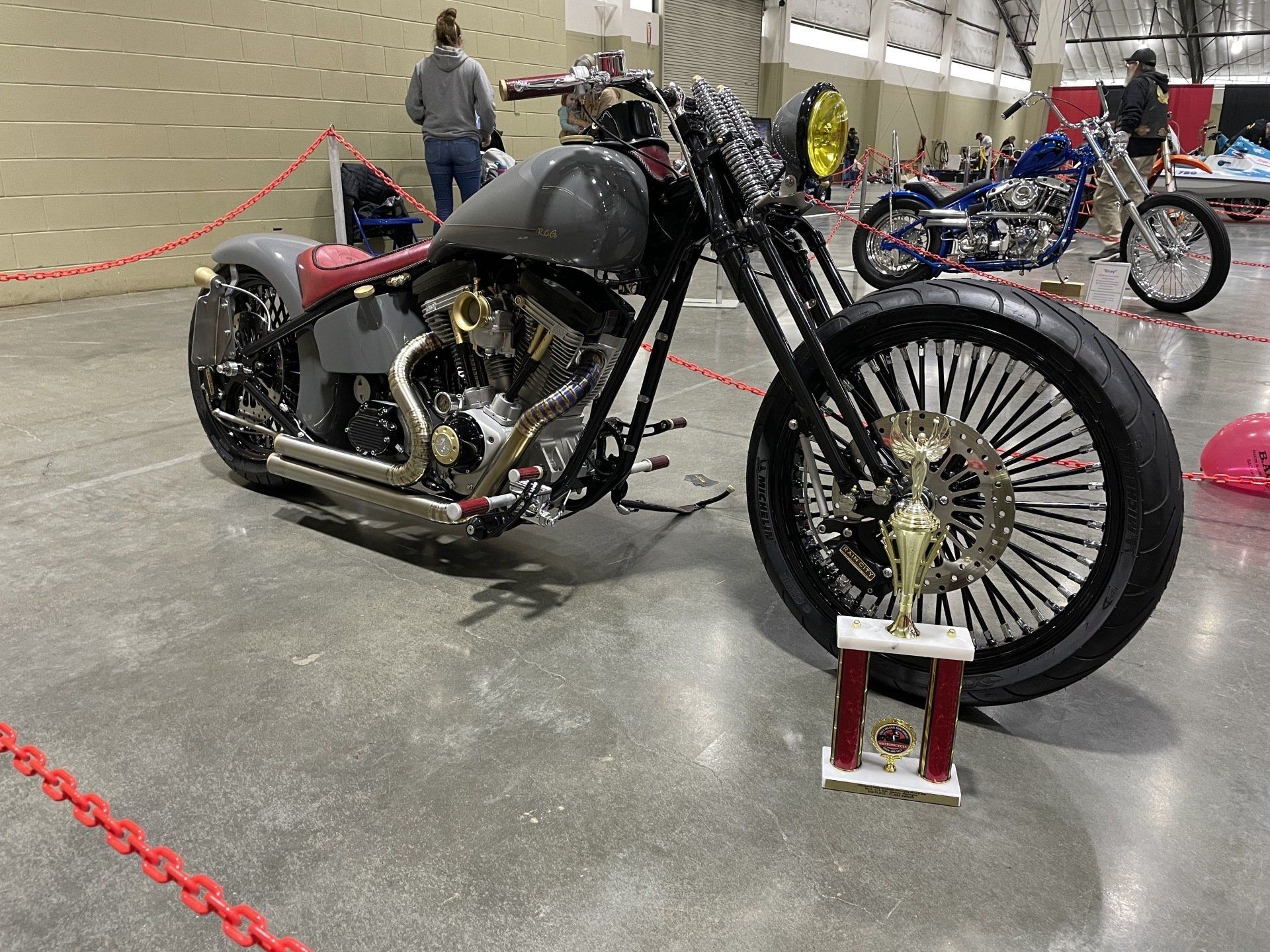 Black custom motorcycle displayed indoors on a concrete floor, with large spoked wheels and a trophy in front.