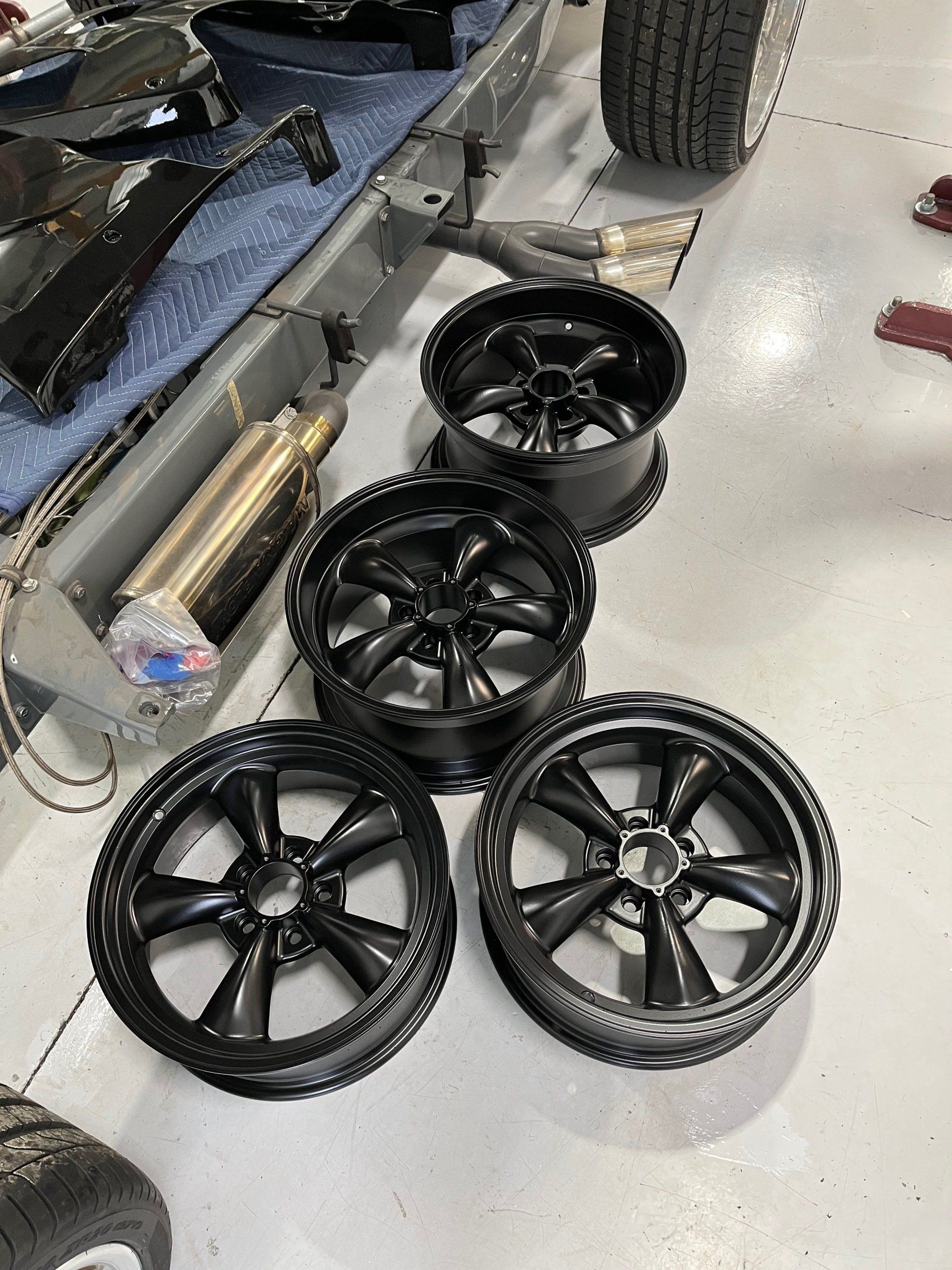 Four glossy black car wheels and tires arranged on a workshop floor, with parts and tools nearby.