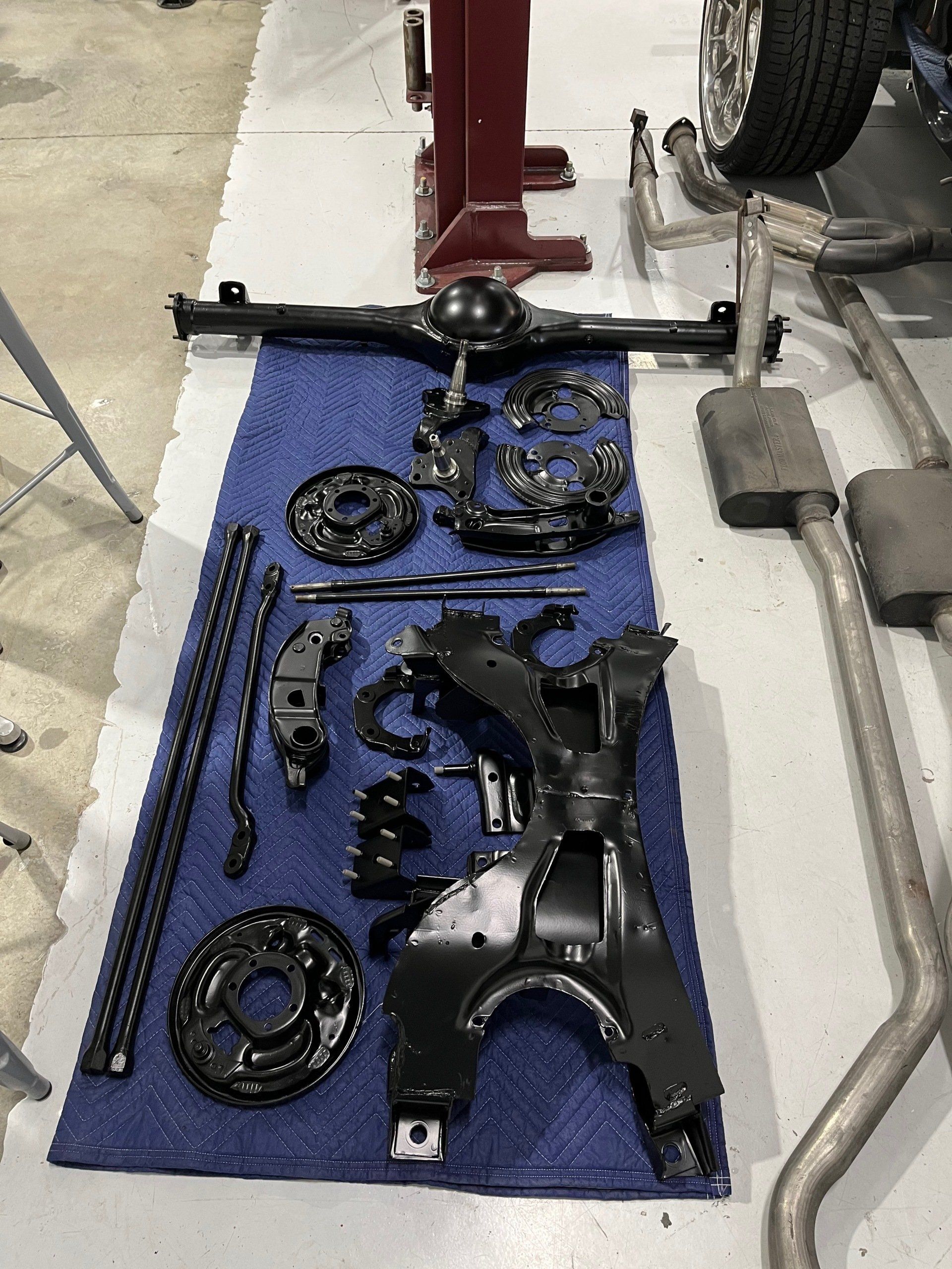 Scattered black automotive suspension parts on a blue mat in a garage, with tools and a vehicle wheel nearby