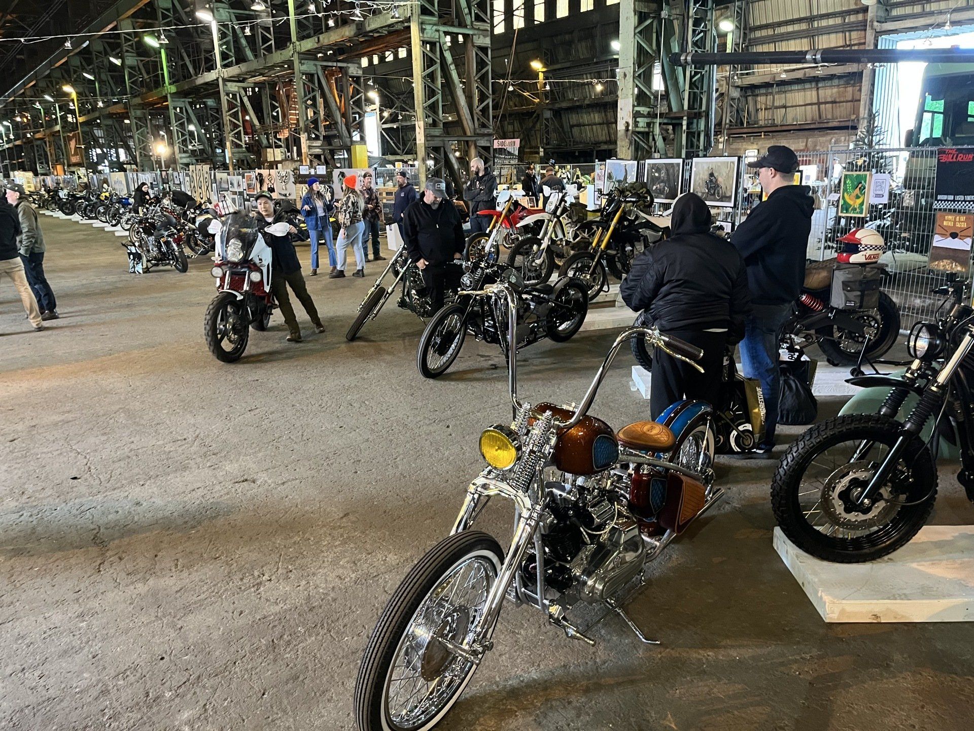 Motorcycles parked in a covered garage with people standing and sitting nearby.
