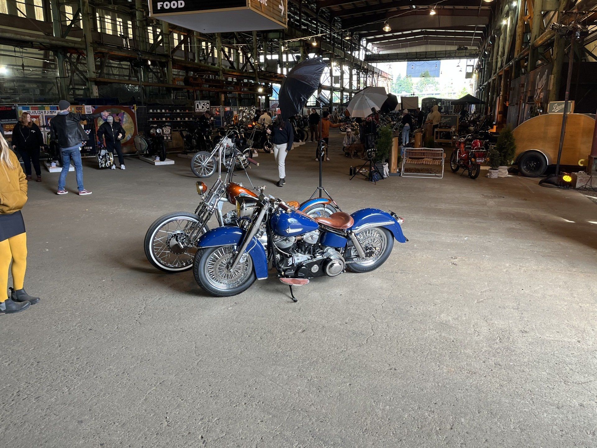 Blue motorcycle parked inside a large industrial warehouse with people and bikes in the background