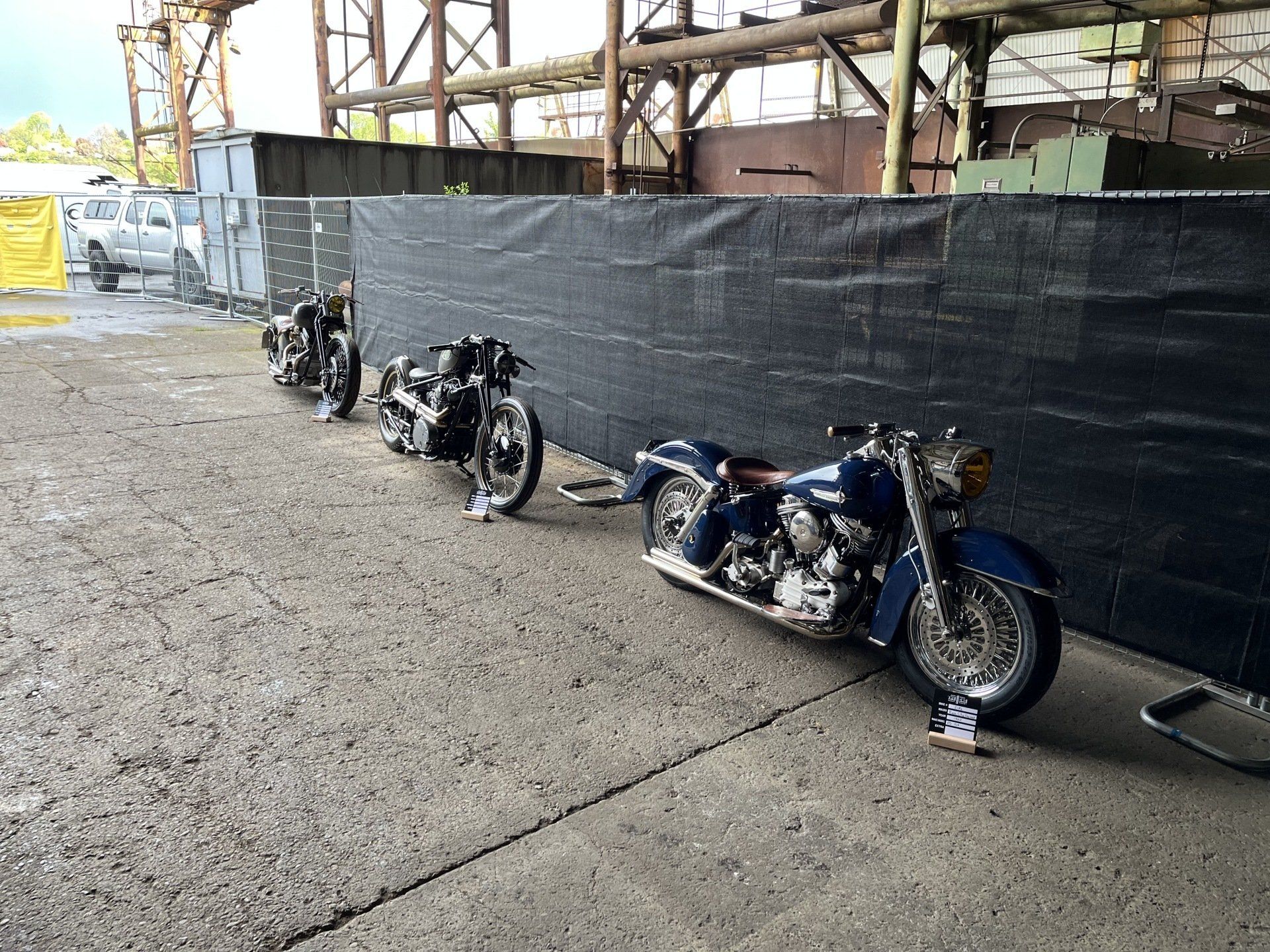 Three custom motorcycles parked beside a black construction fence on a gravel lot.