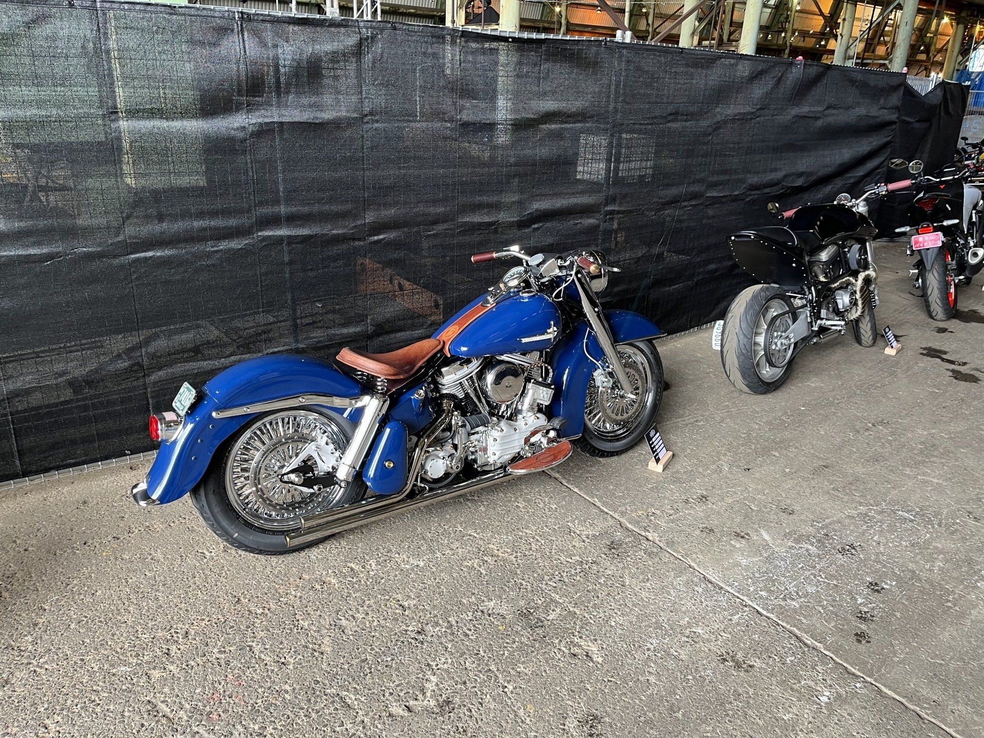 Blue motorcycle parked on a sidewalk beside a black mesh fence, with two other bikes nearby