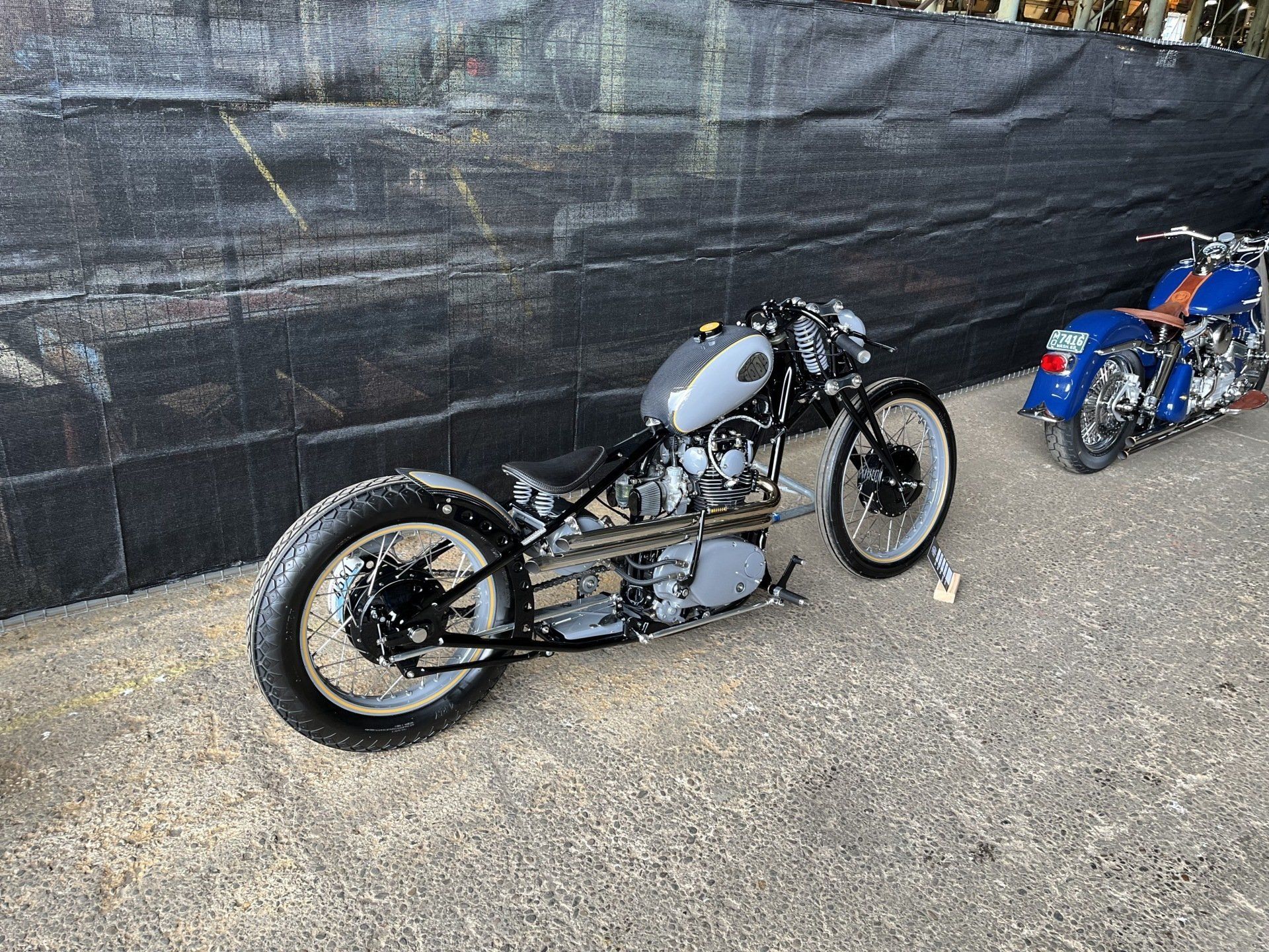 Custom motorcycle with gray tank and black frame parked on gravel beside a dark wall, blue bike in background