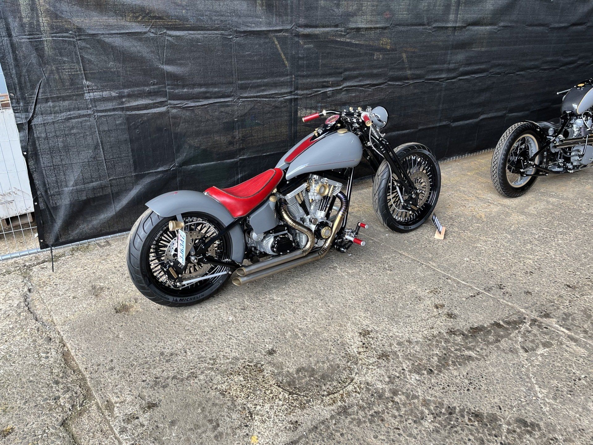 Custom red and gray motorcycle parked on gravel beside a black tarp backdrop