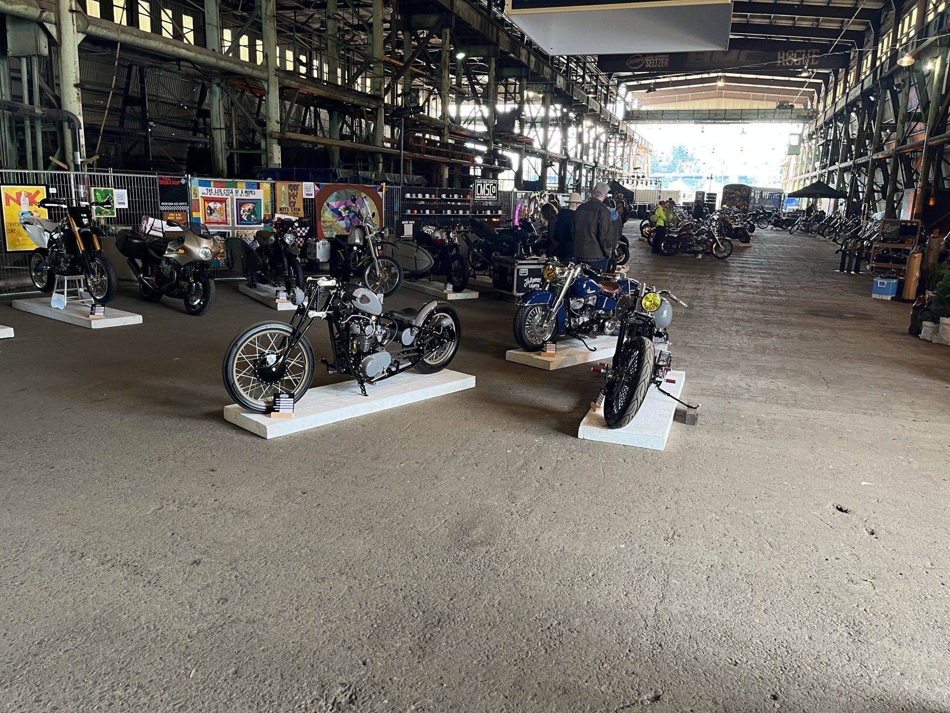 Motorcycles displayed indoors in a large hall with rows of bikes and bright light in the background