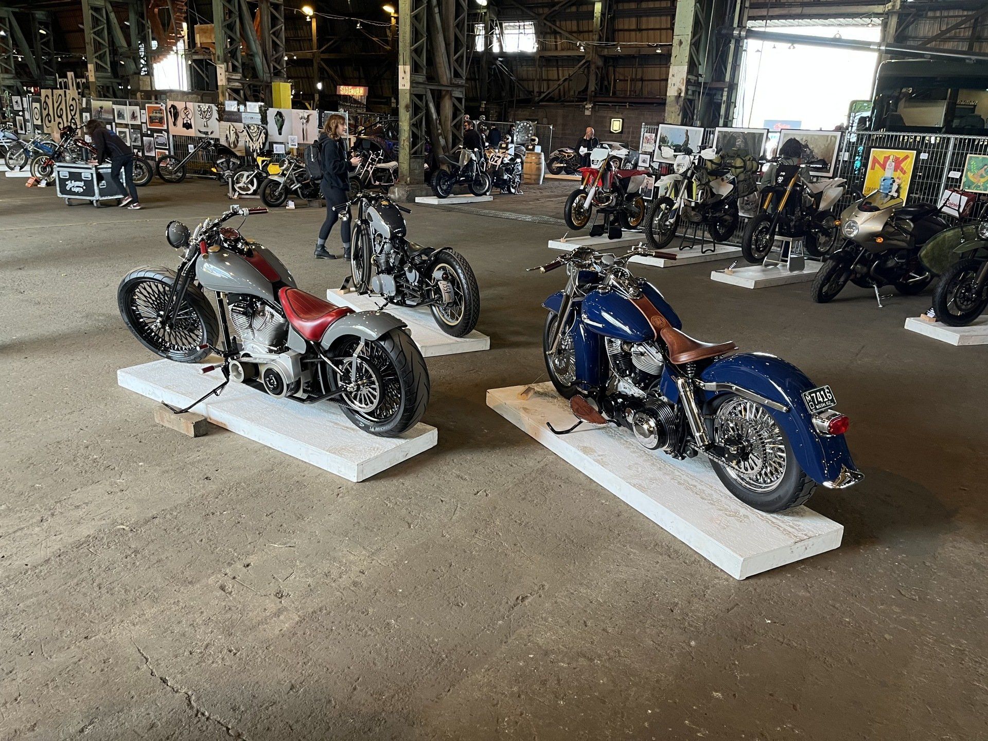 Two custom motorcycles on display indoors, one red and white and one blue, in a workshop or showroom
