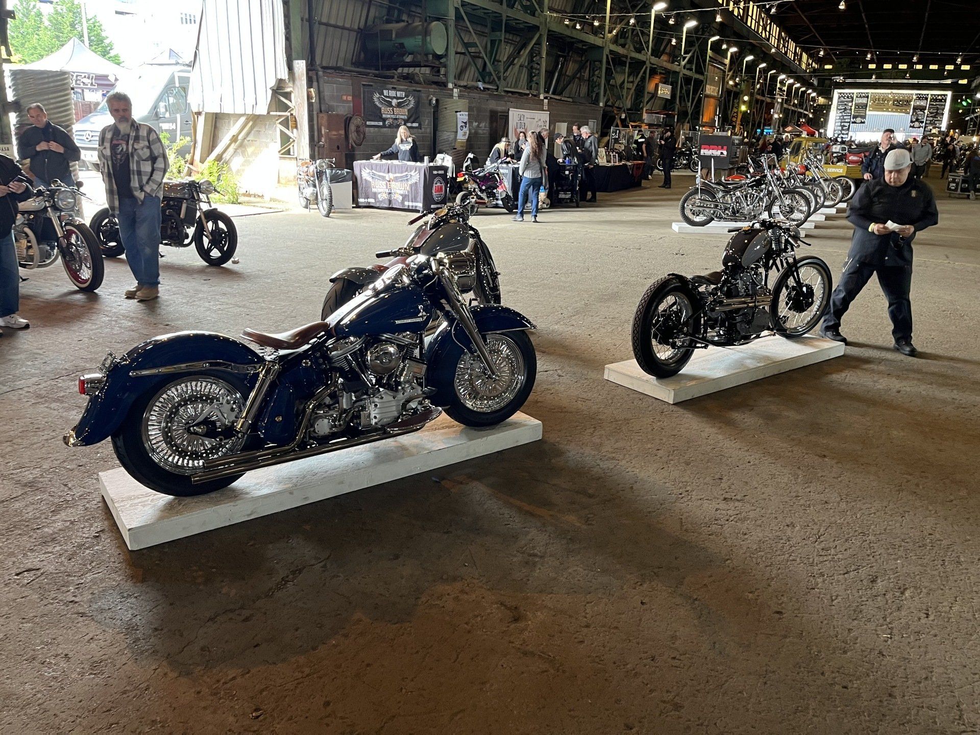 Two custom motorcycles on display in a warehouse-like showroom, one blue and one black on white platforms.