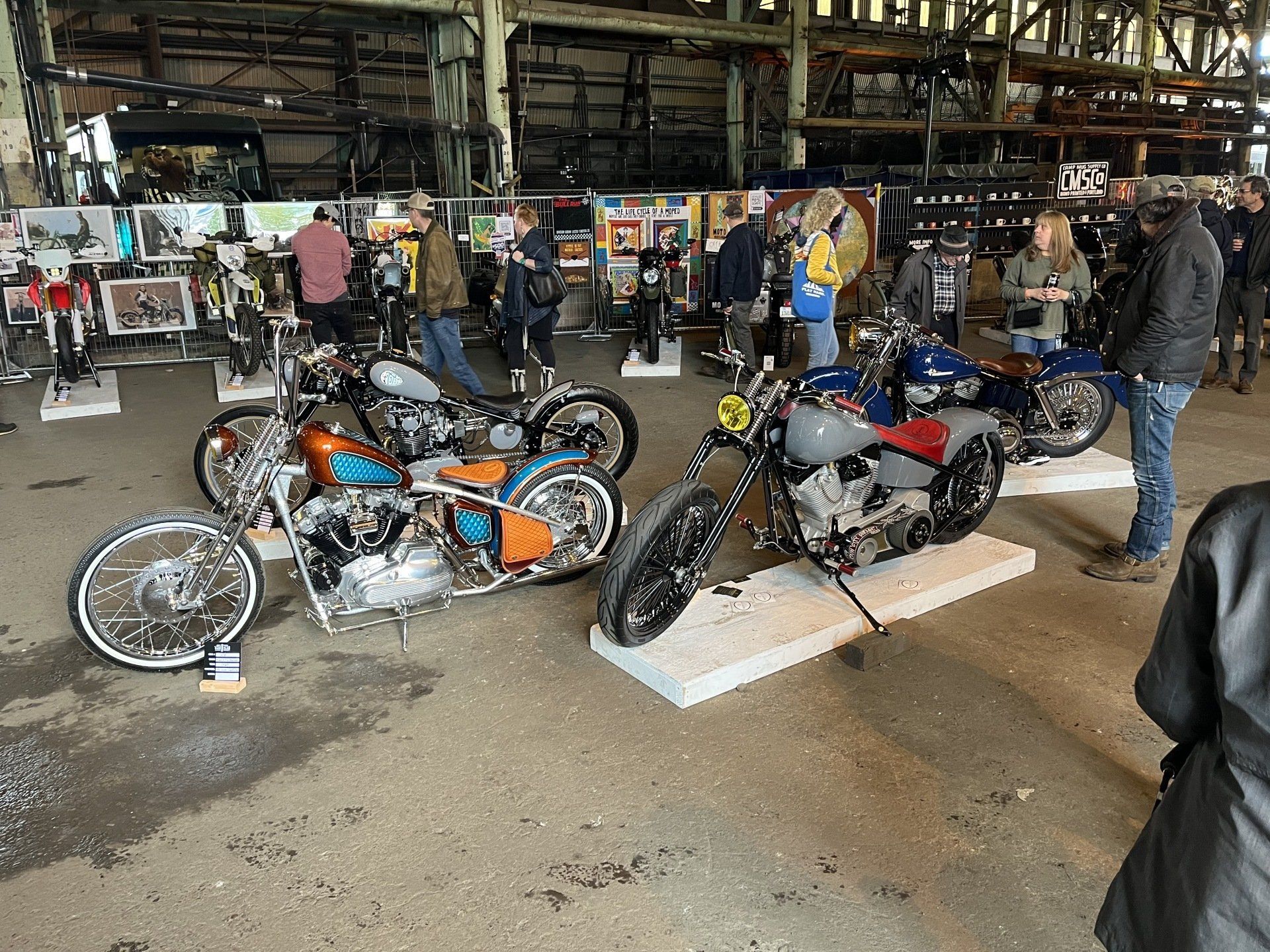 Two custom motorcycles on display in an industrial warehouse, with people browsing nearby