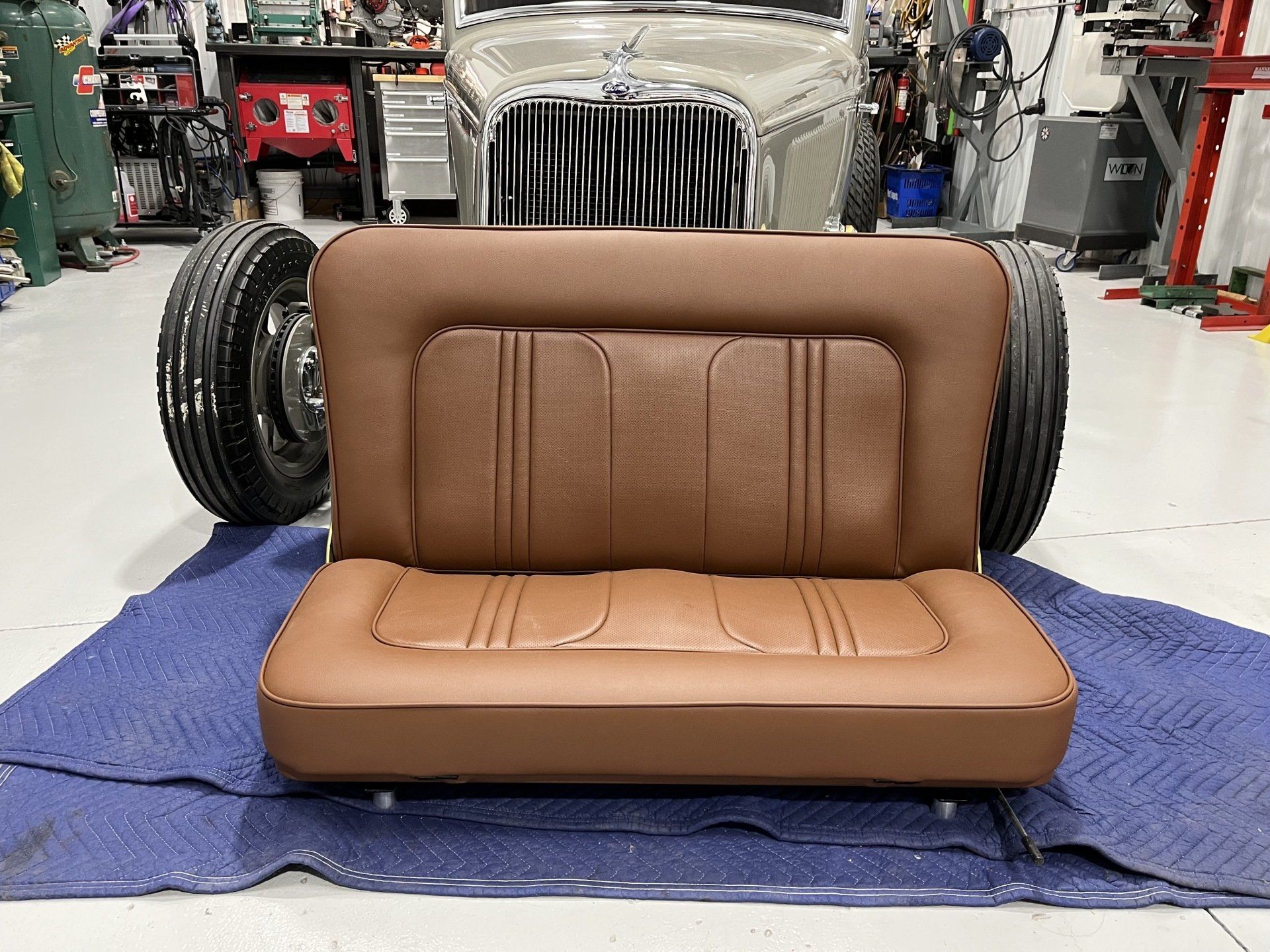 Brown car seat on a blue mat in a garage, with a classic car and wheels in the background
