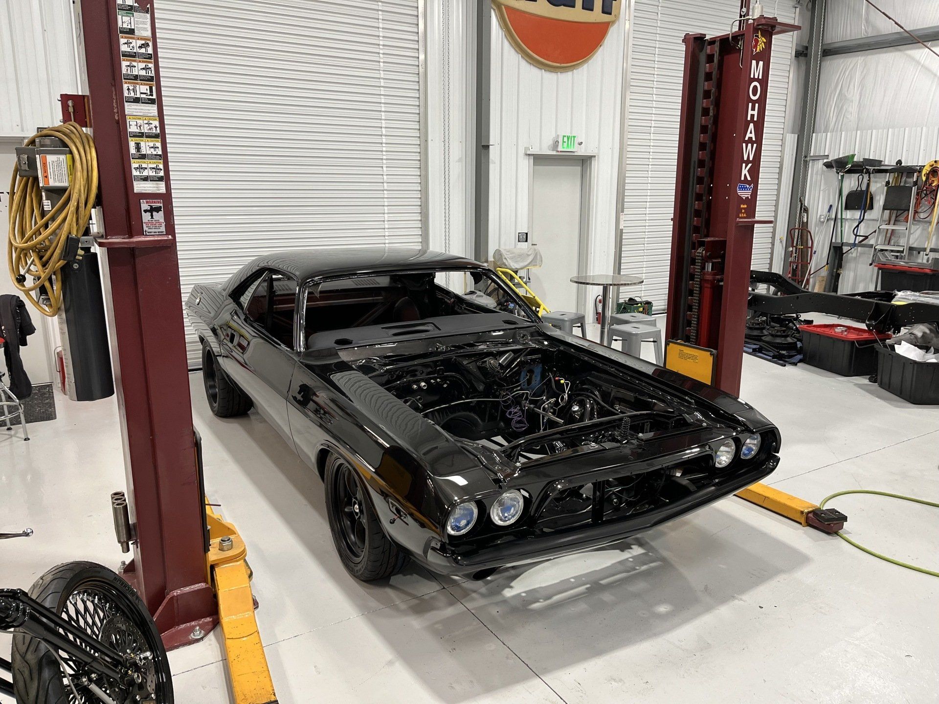 Black classic car shell on a lift in an auto repair shop, with exposed engine bay and missing hood.