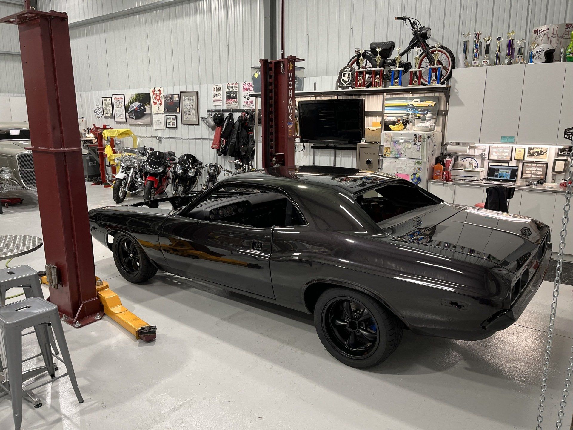 Black classic muscle car in a garage beside a red lift, with motorcycles and tools in the background