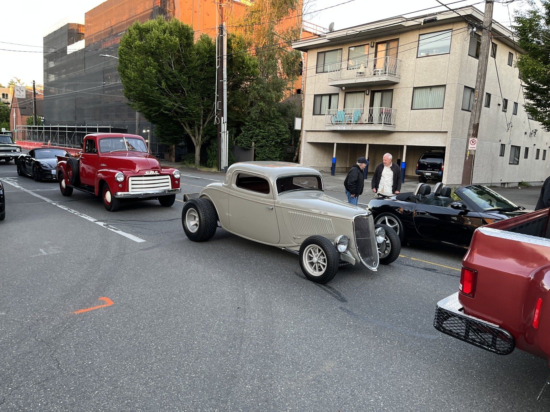 Classic beige hot rod on a city street lined with parked cars and apartment buildings